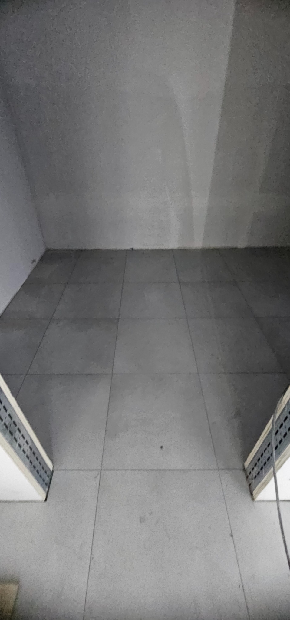 Szare, kwadratowe płytki podłogowe ułożone w pomieszczeniu, widok z góry, z fragmentami listew przypodłogowych po bokach. Minimalistyczny design.