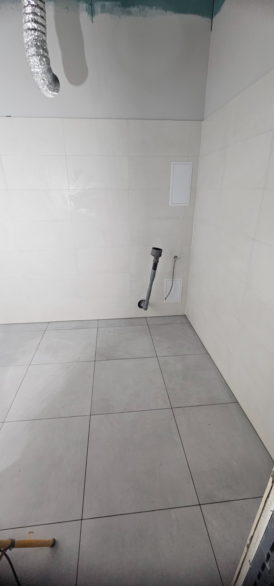 Wykończona łazienka z szarymi płytkami podłogowymi i białymi ściennymi. Widoczny odpływ kanalizacyjny i fragment wentylacji. Ściany częściowo przygotowane do malowania.