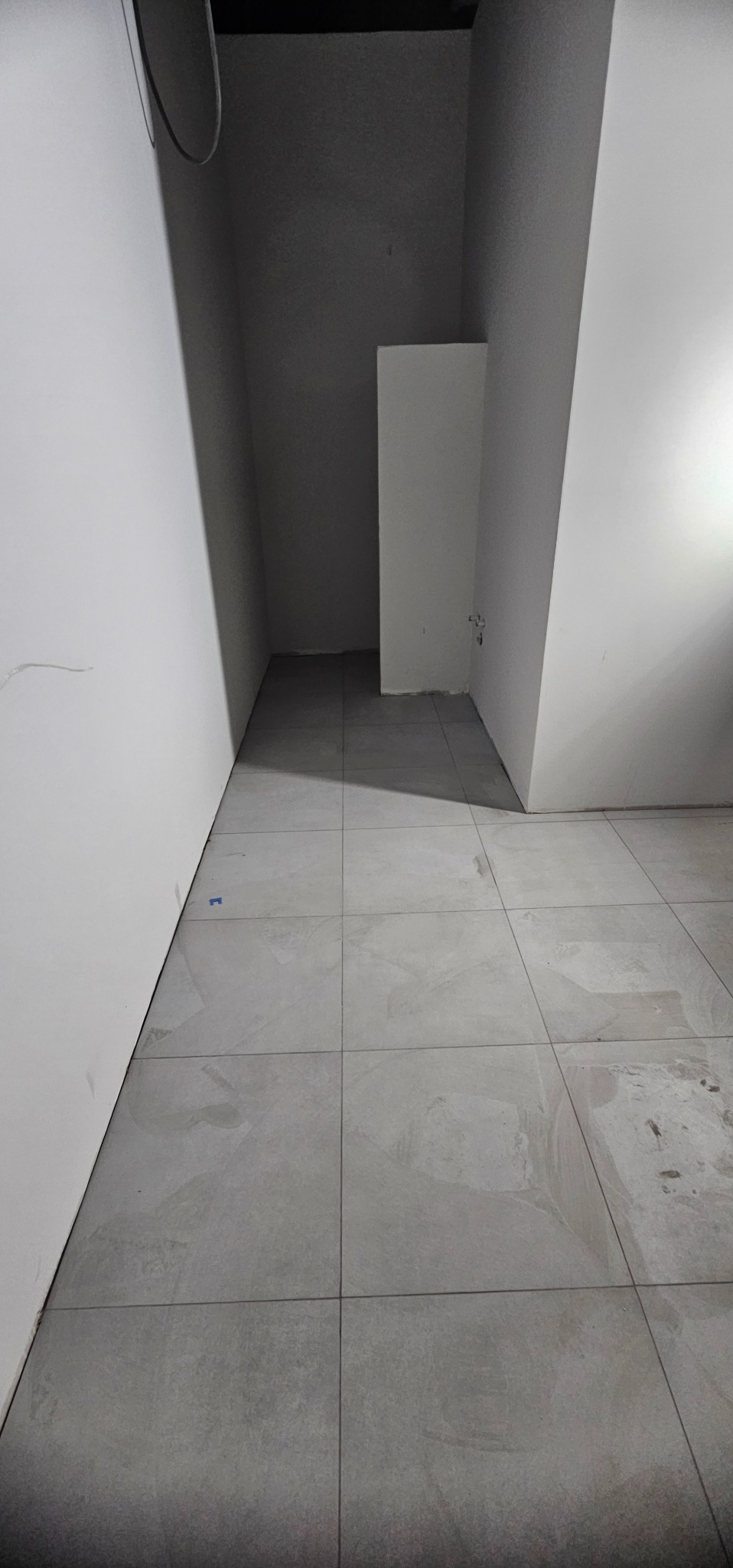 Wykończona podłoga z szarych, kwadratowych płytek w pomieszczeniu z białymi ścianami. Widoczne fugi i fragment instalacji na suficie. Minimalistyczny design.