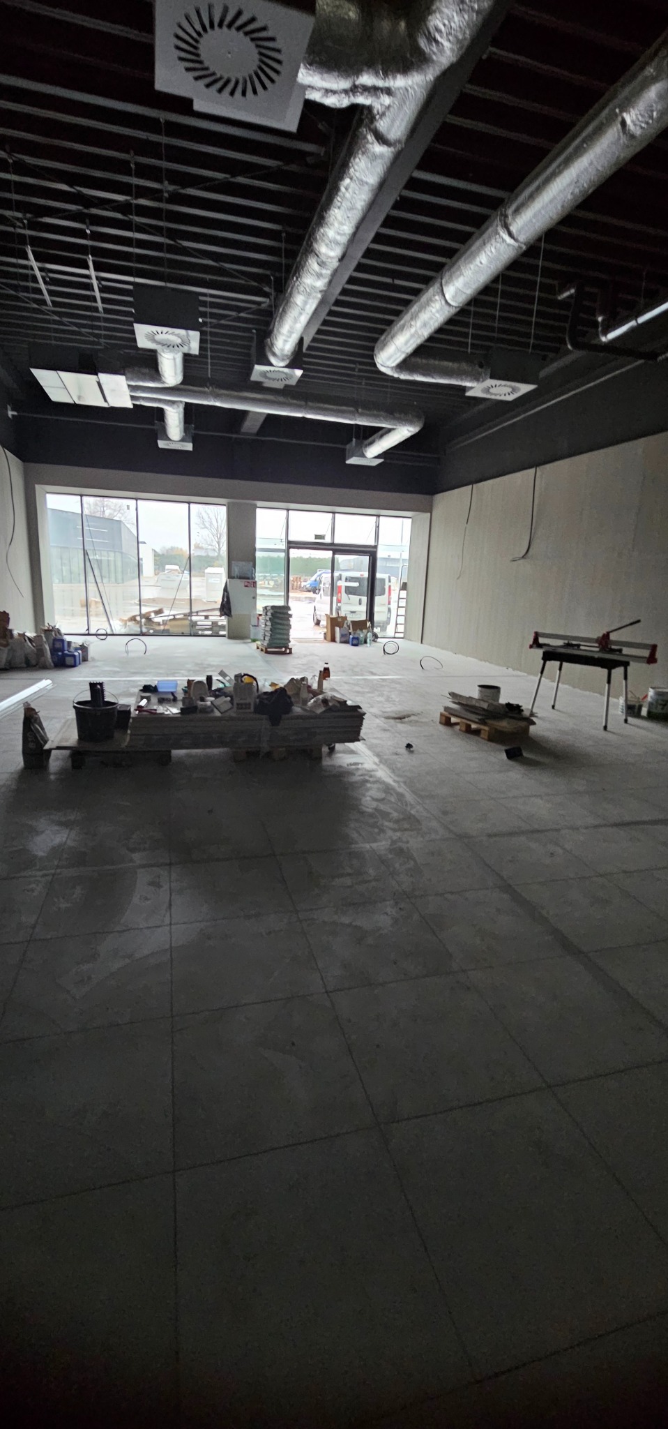 Wnętrze w trakcie prac wykończeniowych: widoczne narzędzia, materiały budowlane i świeżo ułożona podłoga z płytek w dużym pomieszczeniu z oknami i instalacją wentylacyjną.