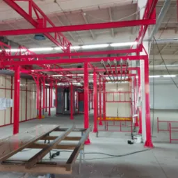 Czerwona konstrukcja stalowa w hali produkcyjnej, prawdopodobnie linia do malowania proszkowego, widoczne elementy zawieszenia i wózki transportowe, w tle szafa sterownicza i dokumentacja techniczna.