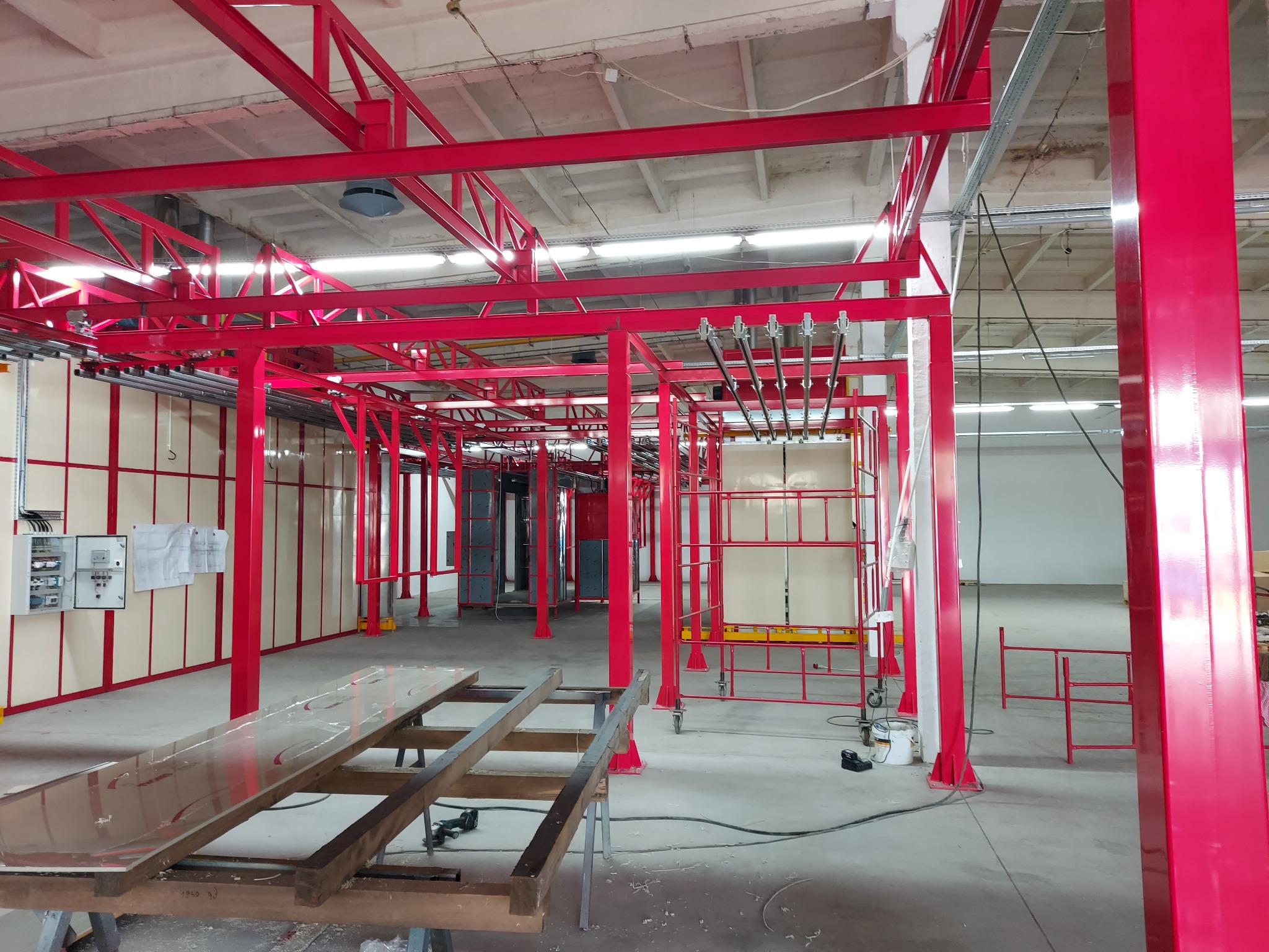 Czerwona konstrukcja stalowa w hali produkcyjnej, prawdopodobnie linia do malowania proszkowego, widoczne elementy zawieszenia i wózki transportowe, w tle szafa sterownicza i dokumentacja techniczna.