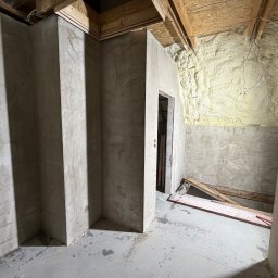 M-KU Wykończenia - Surowe wykończenie piwnicy: ściany z szarego betonu, izolacja piankowa na suficie, widoczne drewniane elementy konstrukcyjne. Wnętrze w trakcie prac wykończeniowych.