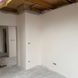 M-KU Wykończenia - Wnętrze po szpachlowaniu ścian, widoczne gładkie powierzchnie przygotowane do malowania. Otwory na gniazdka elektryczne. Surowy stan podłogi i drewniana konstrukcja sufitu.