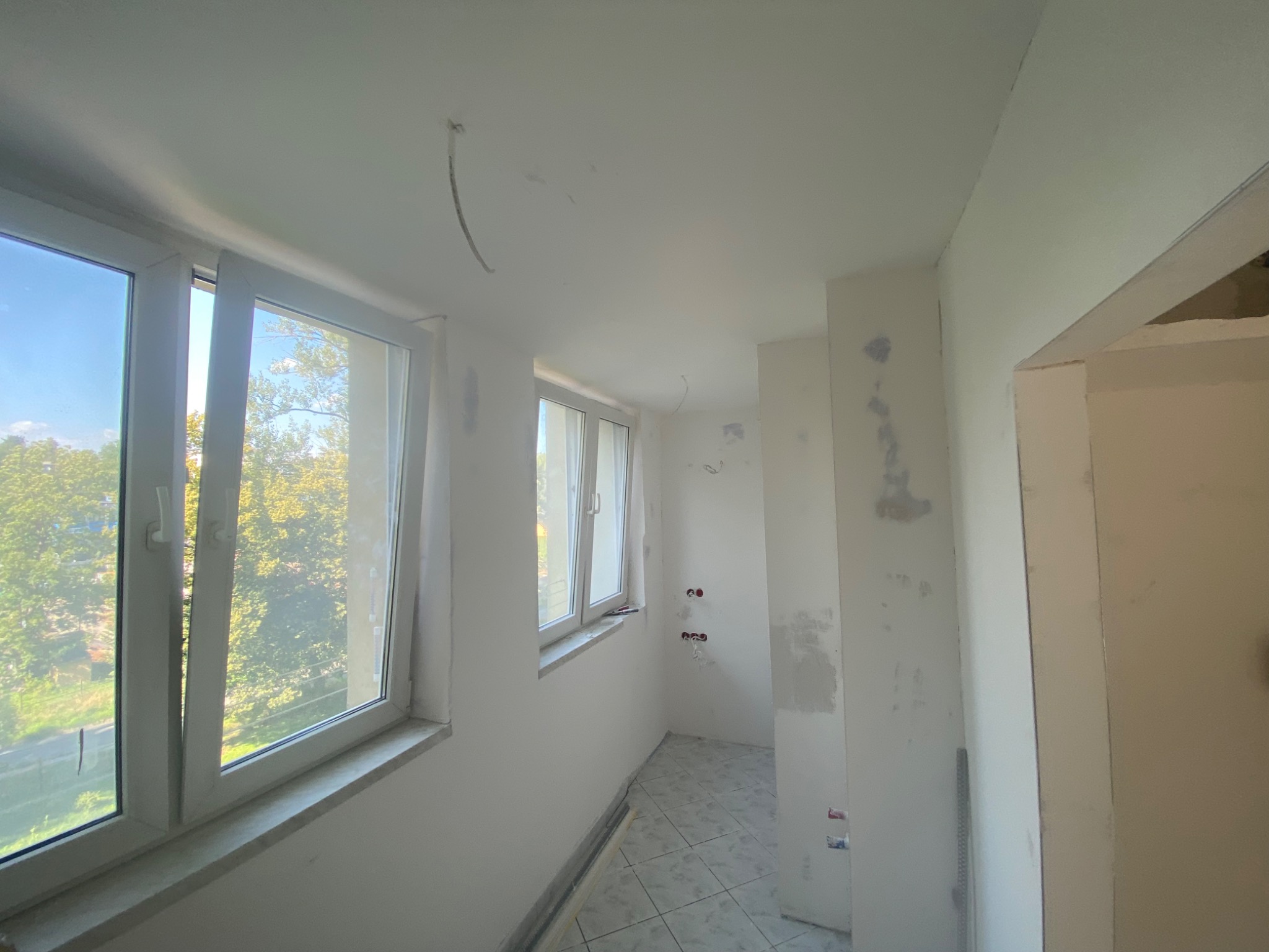 Wnętrze w trakcie remontu: ściany przygotowane do malowania, widoczne ślady szpachlowania, okna z widokiem na zieleń. Sufit z wystającymi kablami.