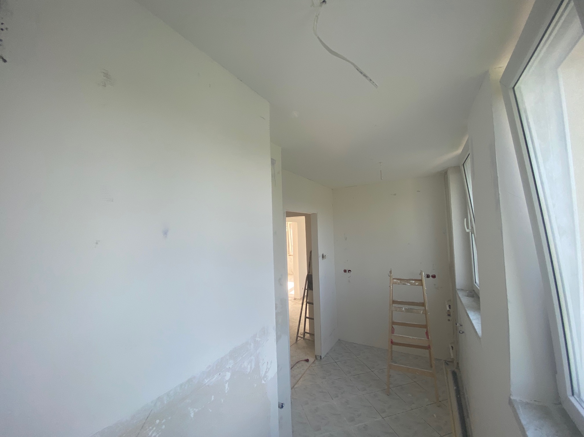 Wnętrze w trakcie remontu: białe ściany, częściowo wykończone gładzie, drabina oparta o ścianę, widoczne przewody elektryczne, okno z białymi ramami.