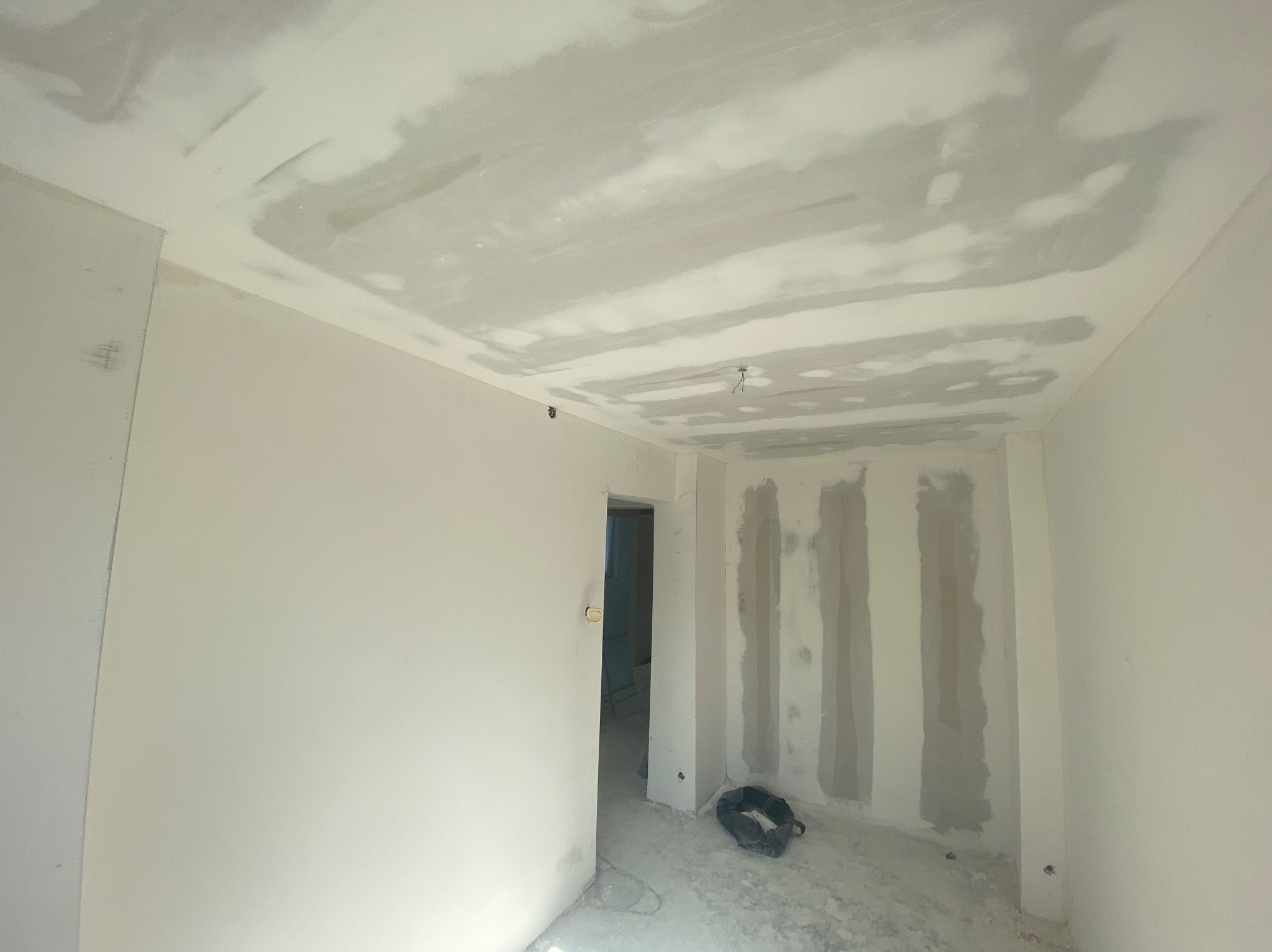 Pokój w trakcie prac wykończeniowych: ściany i sufit po szpachlowaniu, widoczne ślady po łatach. Drzwi otwarte do kolejnego pomieszczenia, narzędzia na podłodze.