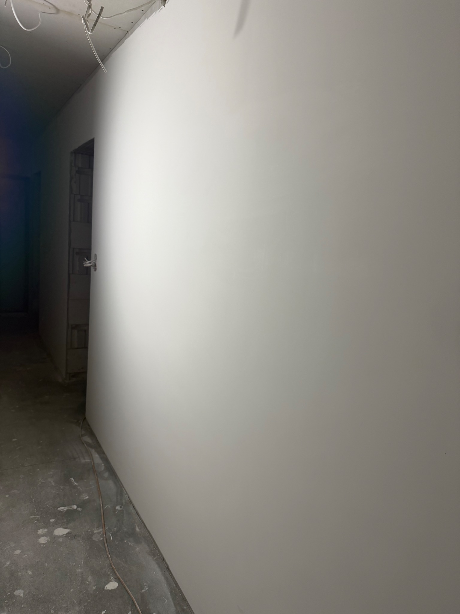 Gładka, biała ściana po szpachlowaniu w surowym wnętrzu. Widoczne kable wystające z sufitu i fragment ściany z pustaków w przejściu. Minimalistyczne ujęcie.