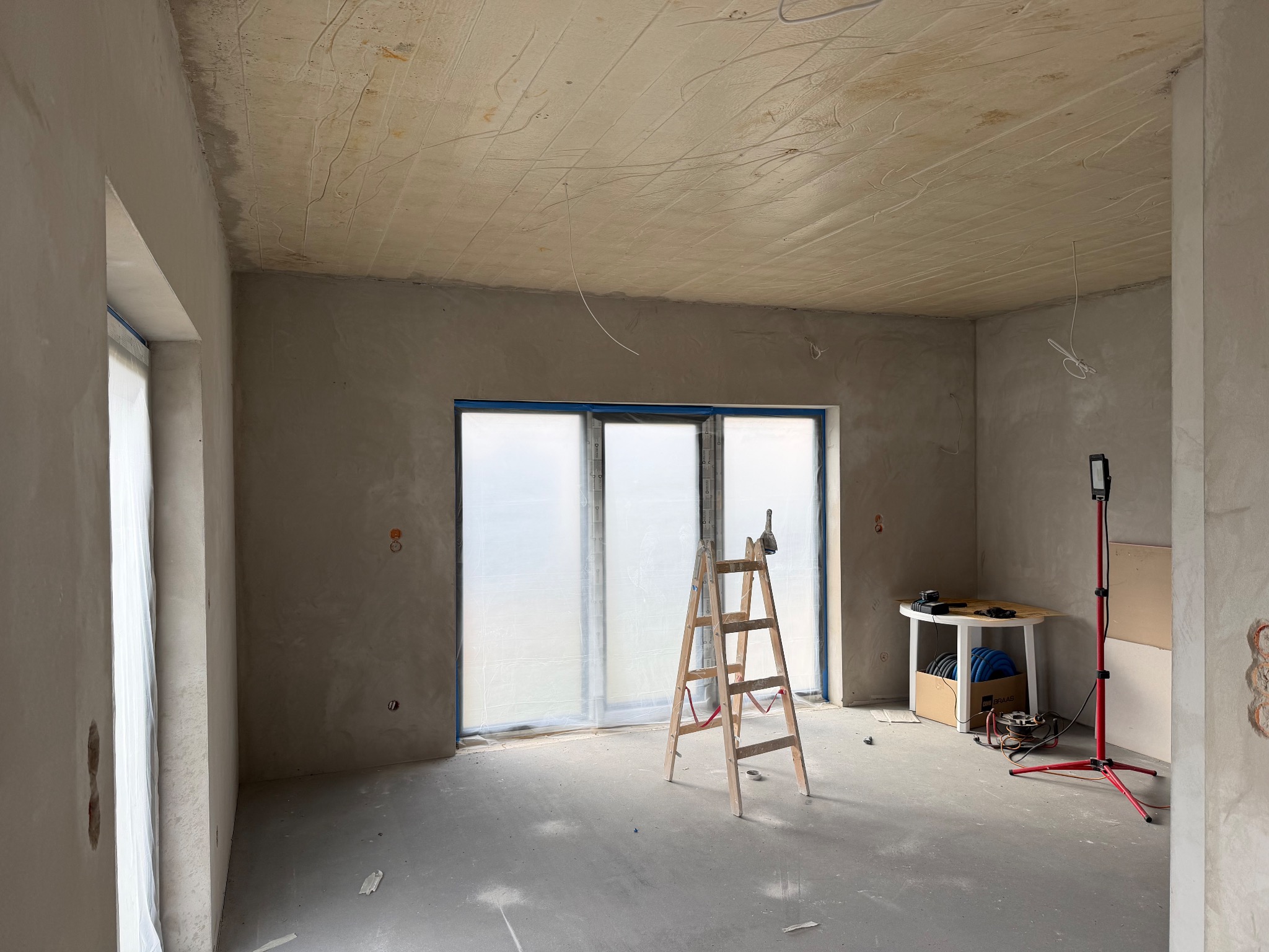 Pokój w trakcie remontu: ściany i sufit po szpachlowaniu, okna zabezpieczone folią, drabina, lampa budowlana i stół z narzędziami. Widoczne ślady po instalacjach.