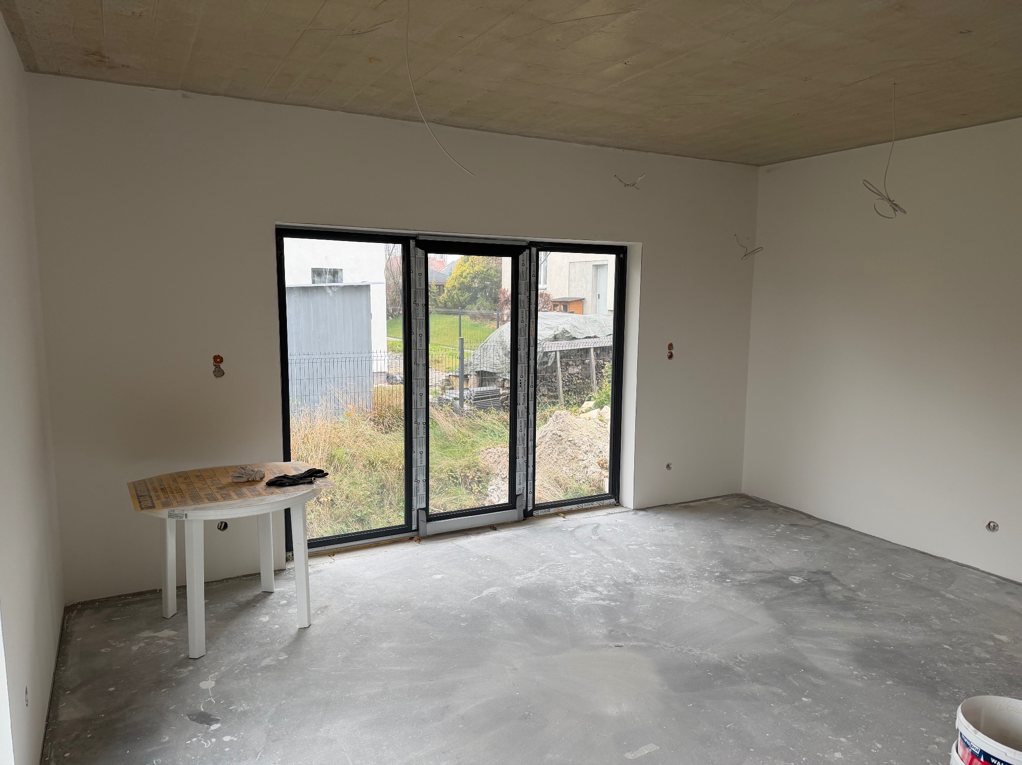Pokój w trakcie remontu: białe ściany, surowy betonowy sufit, duże okno z widokiem na zewnątrz. Na podłodze ślady po wylewce, w rogu stoi biały stolik z rękawicami.