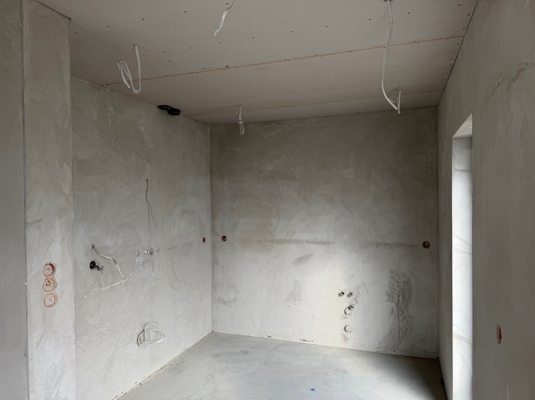 Surowe wnętrze pokoju w trakcie remontu: ściany pokryte gładzią, widoczne instalacje elektryczne, sufit z płyt karton-gips, przygotowane do dalszych prac wykończeniowych.
