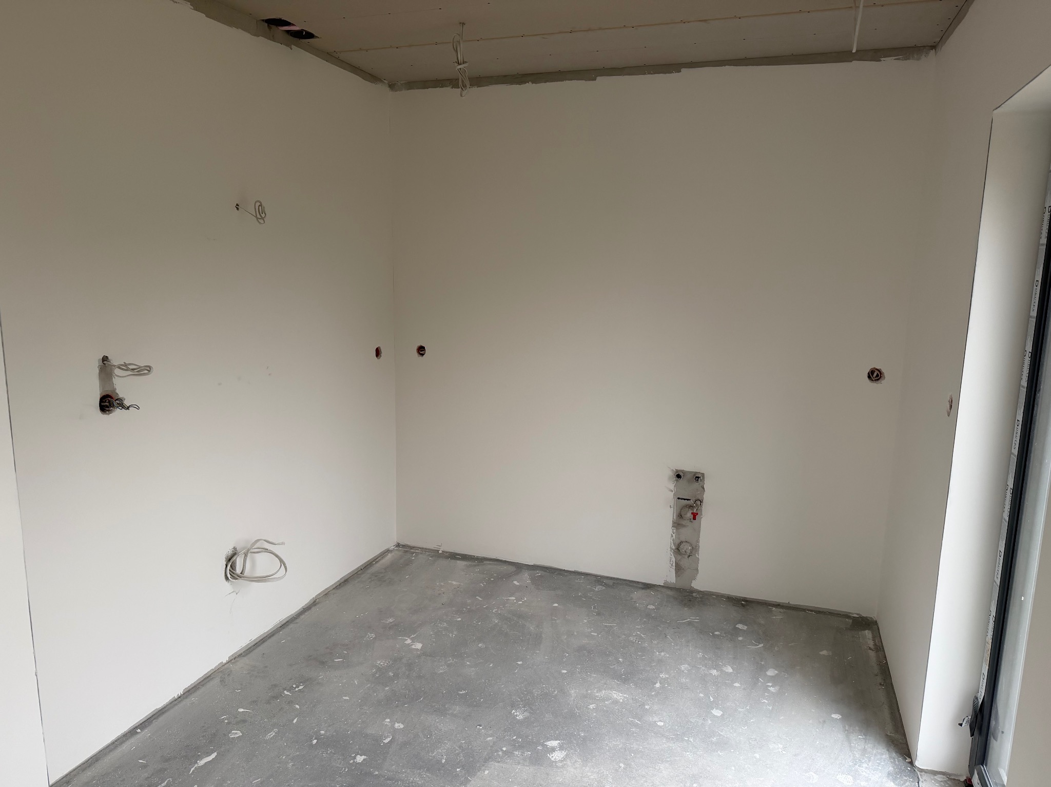 Wnętrze w stanie deweloperskim: gładkie, białe ściany, surowy beton na podłodze, widoczne instalacje elektryczne i wodne. Przygotowanie do wykończenia.