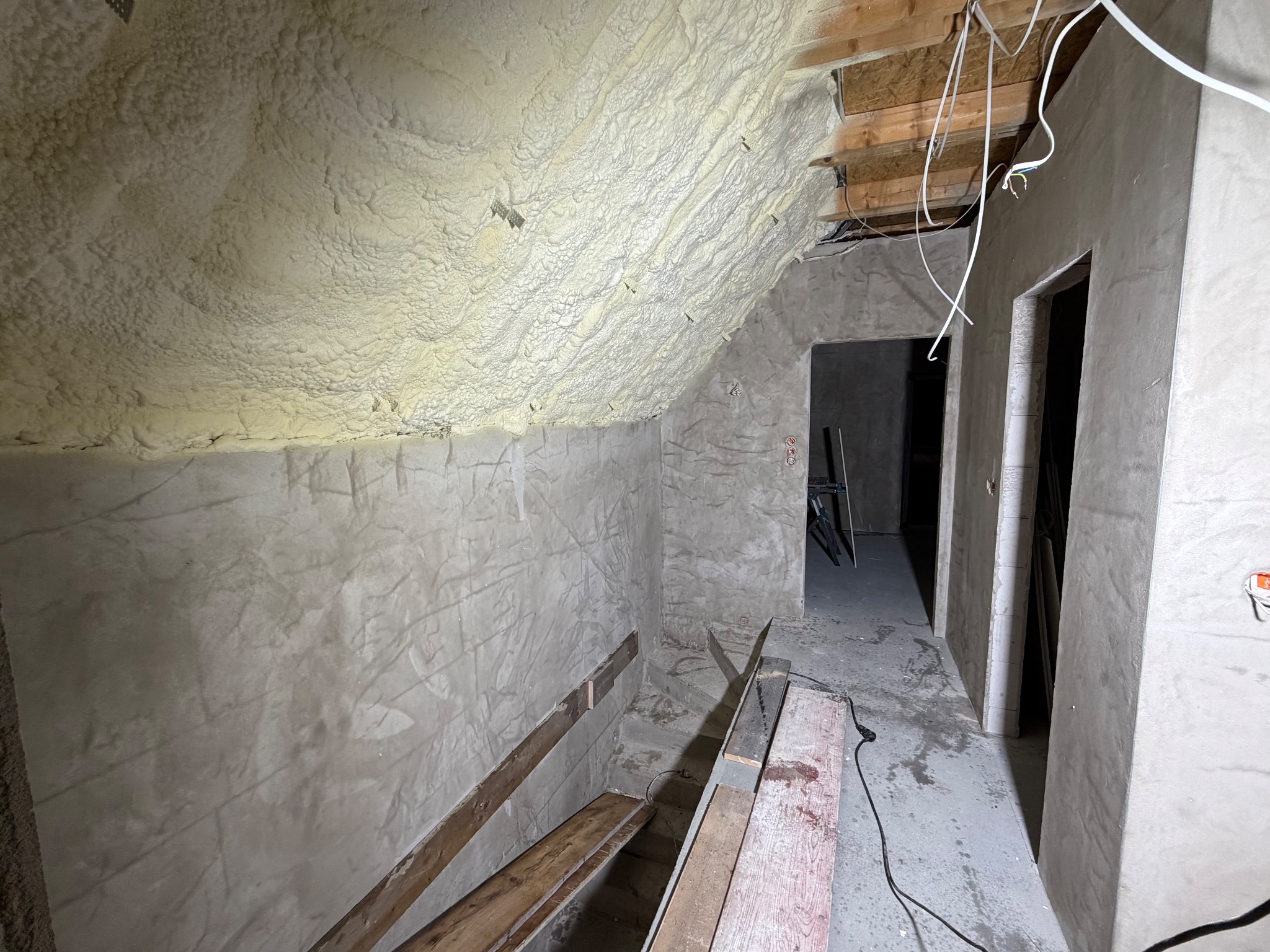 Poddasze w trakcie prac wykończeniowych: ściany pokryte szpachlą, izolacja piankowa na skosach, widoczne przewody elektryczne i elementy konstrukcyjne. Surowy stan wnętrza.