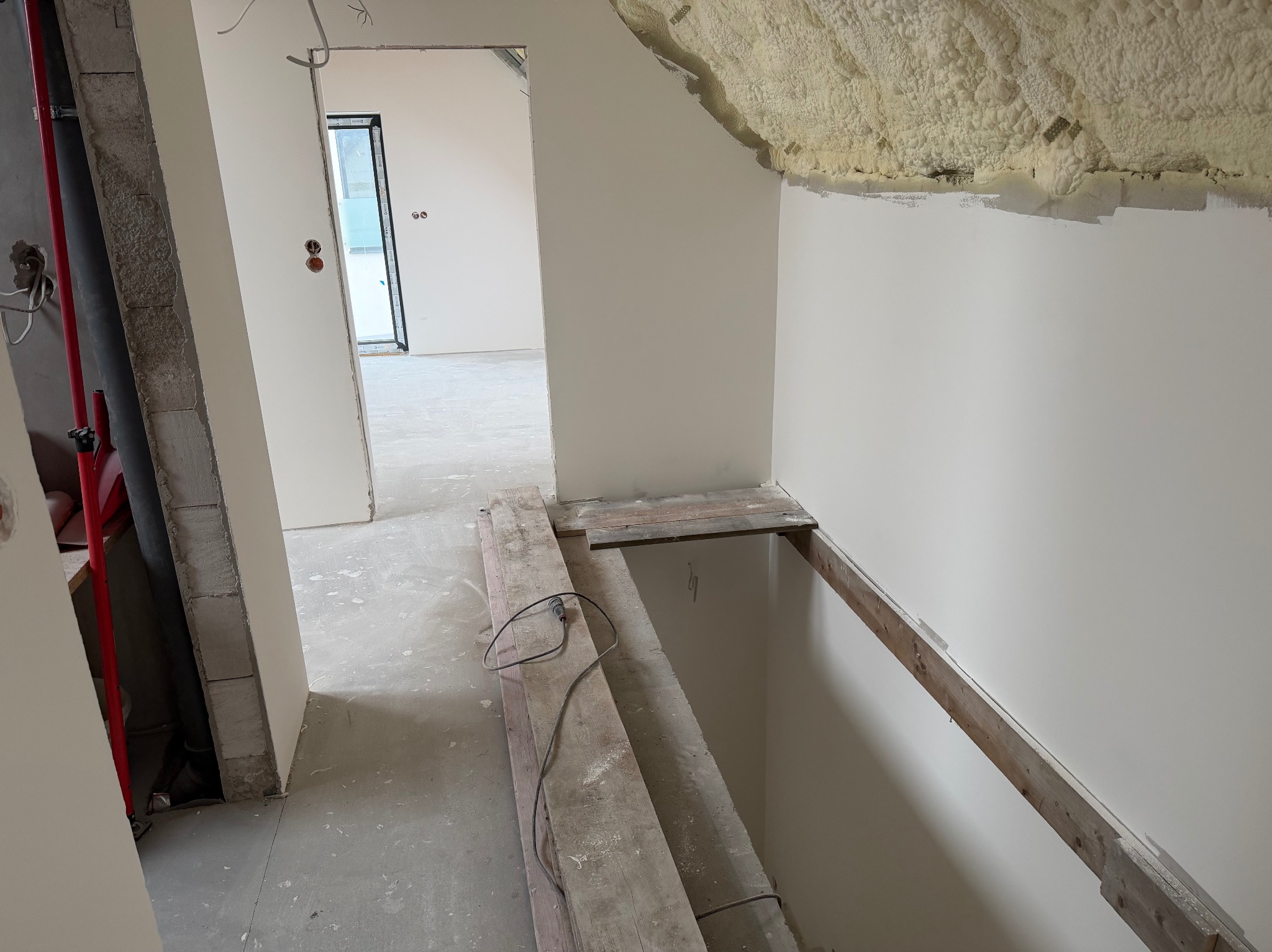 Poddasze w trakcie wykańczania: ocieplony strop pianką, świeżo pomalowane ściany na biało, widoczne deski podłogowe i instalacje. W tle otwarte drzwi do kolejnego pomieszczenia.