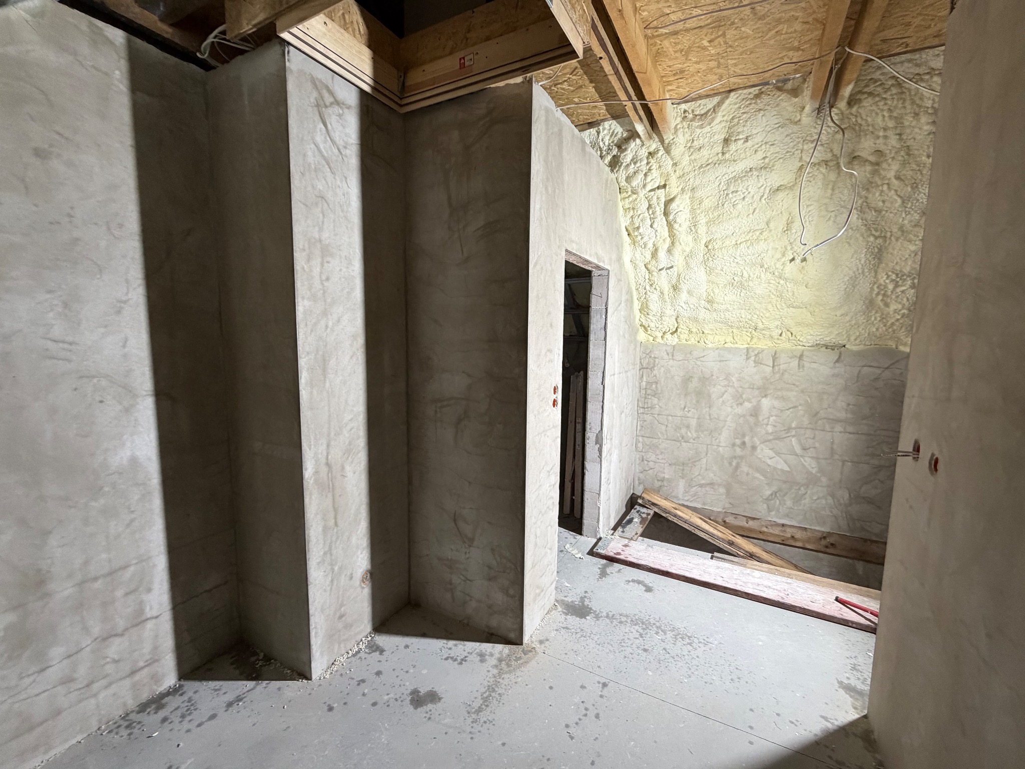 Surowe wykończenie piwnicy: ściany z szarego betonu, izolacja piankowa na suficie, widoczne drewniane elementy konstrukcyjne. Wnętrze w trakcie prac wykończeniowych.