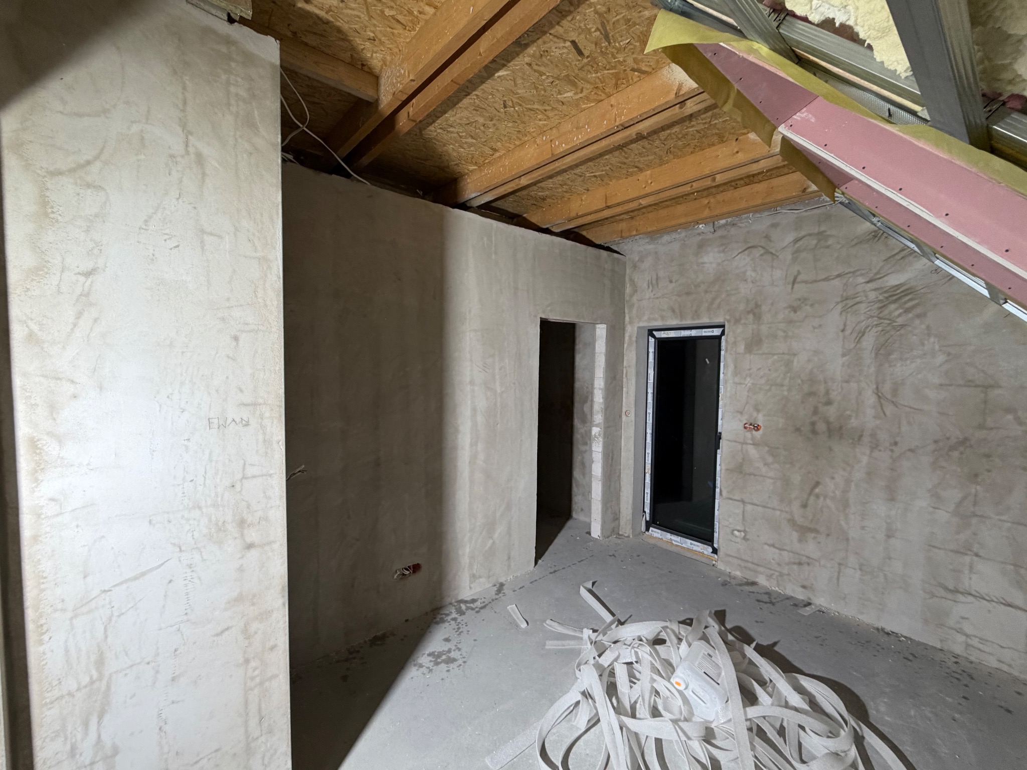 Surowe wnętrze poddasza ze ścianami po szpachlowaniu, widoczne elementy konstrukcji dachu i resztki taśmy malarskiej na podłodze. Przygotowanie do dalszych prac wykończeniowych.