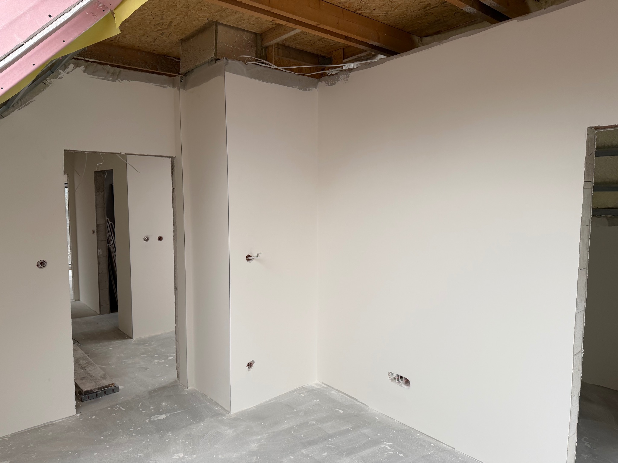 Wnętrze po szpachlowaniu ścian, widoczne gładkie powierzchnie przygotowane do malowania. Otwory na gniazdka elektryczne. Surowy stan podłogi i drewniana konstrukcja sufitu.