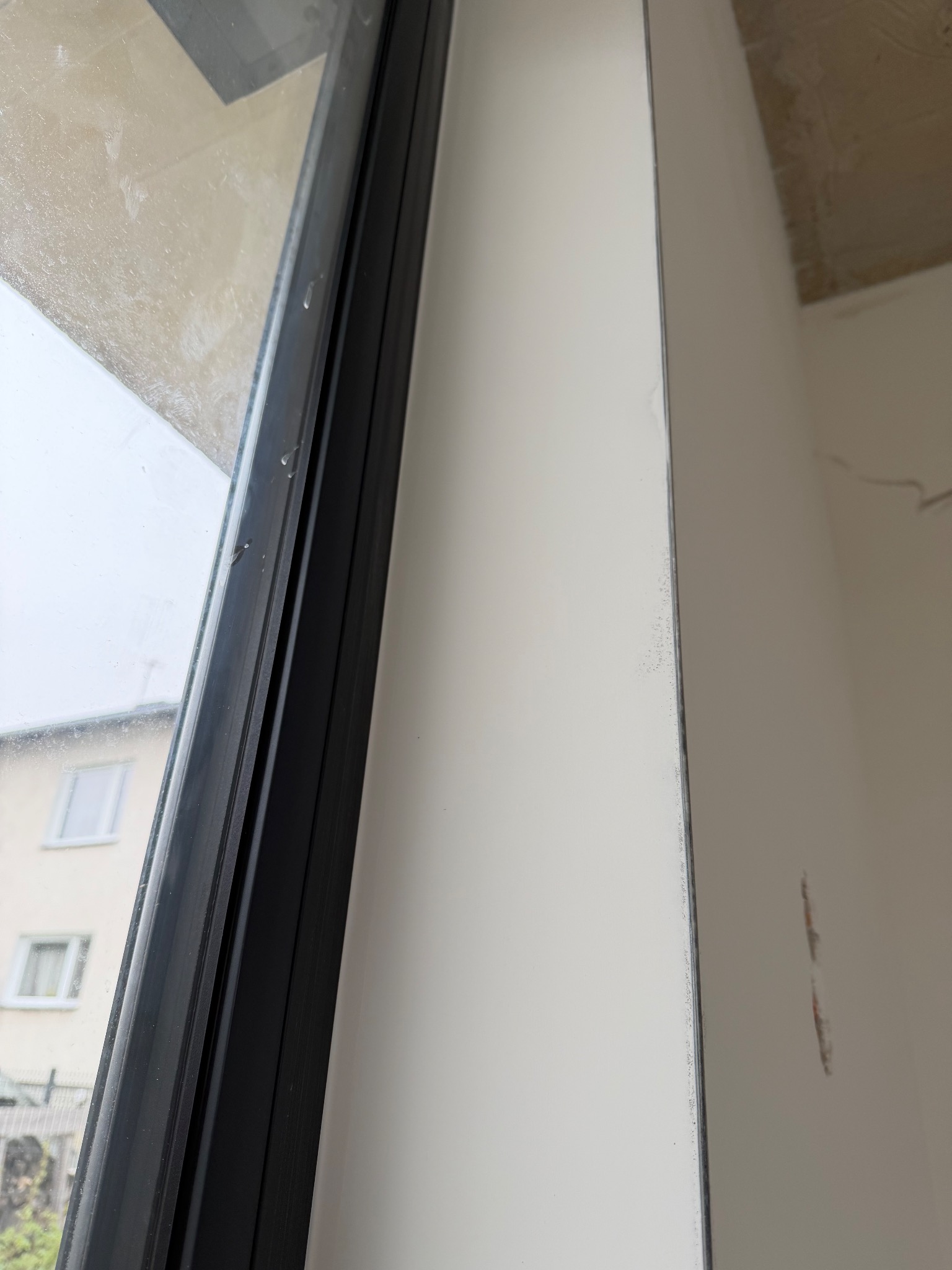 Biała ściana po szpachlowaniu i malowaniu, widoczna przy oknie z czarną ramą. Fragment surowego sufitu w rogu. Świeżo wykończone wnętrze.