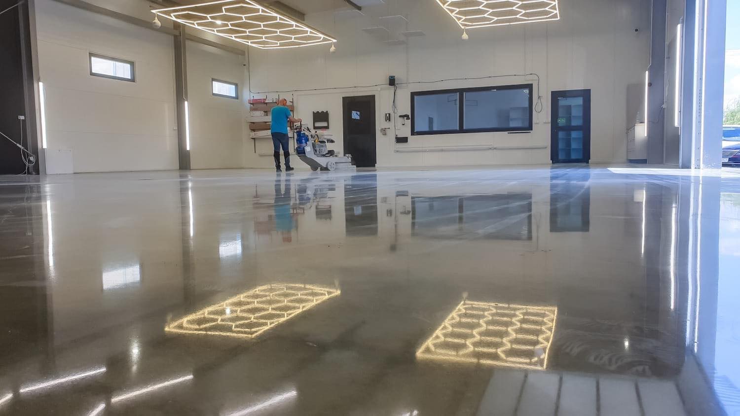 Pracownik w studio detailingowym poleruje posadzkę maszyną do polerowania, widoczne odbicia lamp sufitowych na błyszczącej, zabezpieczonej powierzchni.