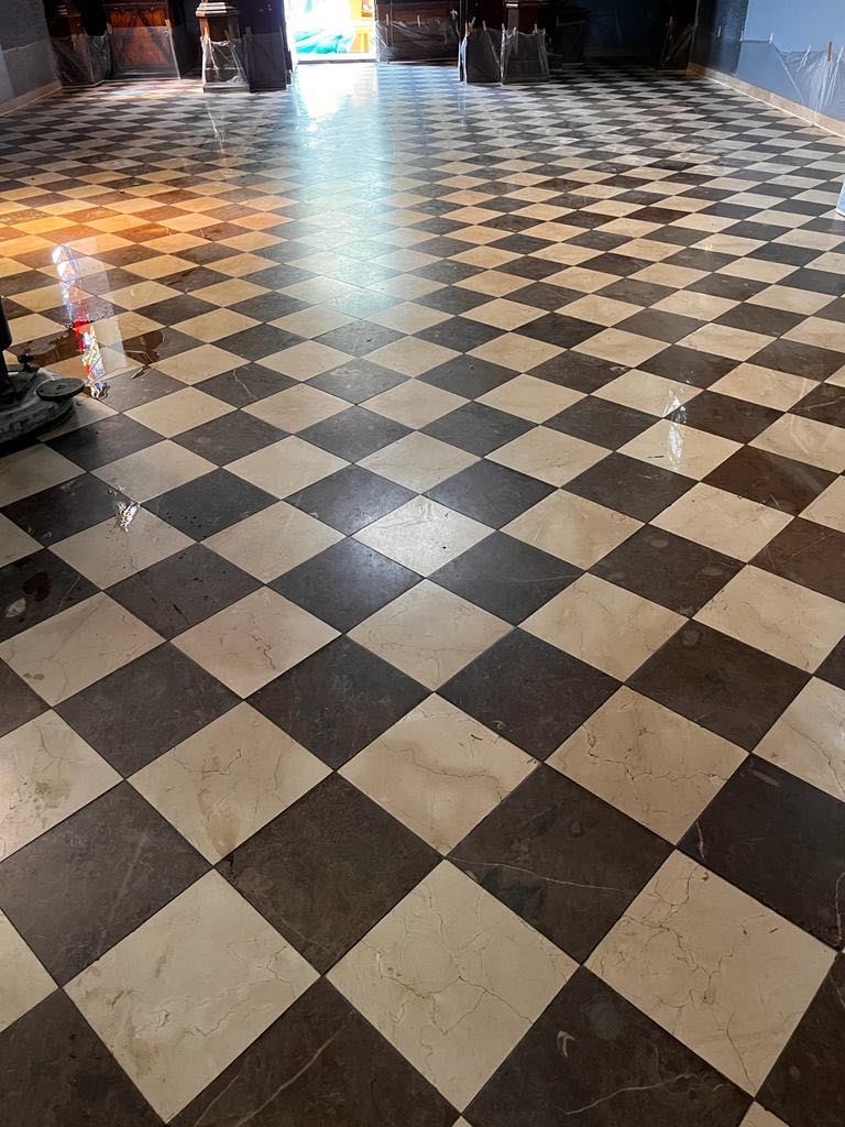 Polerowana posadzka w parafii z czarno-białych, marmurowych kwadratów, częściowo mokra po czyszczeniu, z widocznym fragmentem maszyny czyszczącej po lewej stronie.