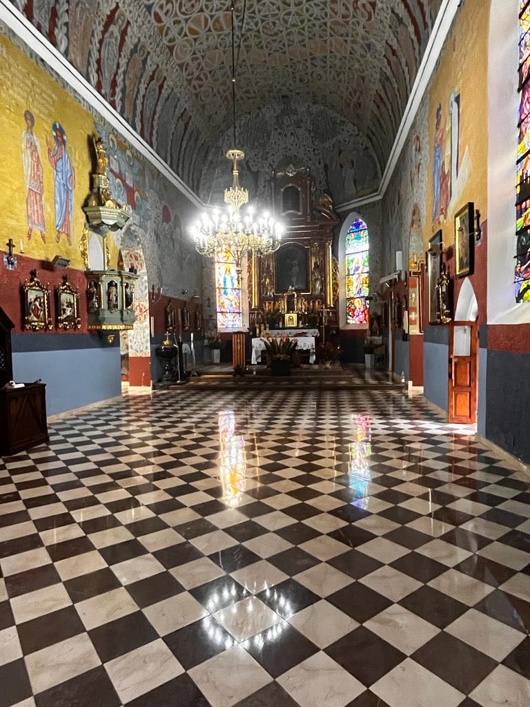 Wnętrze kościoła z polerowaną, czarno-białą posadzką w szachownicę, odbijającą światło z okien i żyrandola, ściany zdobione malowidłami i rzeźbami.