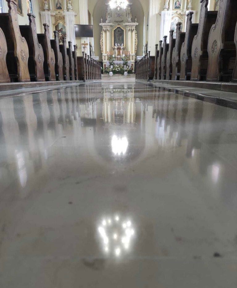 Polerowana posadzka marmurowa odbija światło wewnątrz kościoła, widoczne drewniane ławki po bokach i ołtarz w oddali.