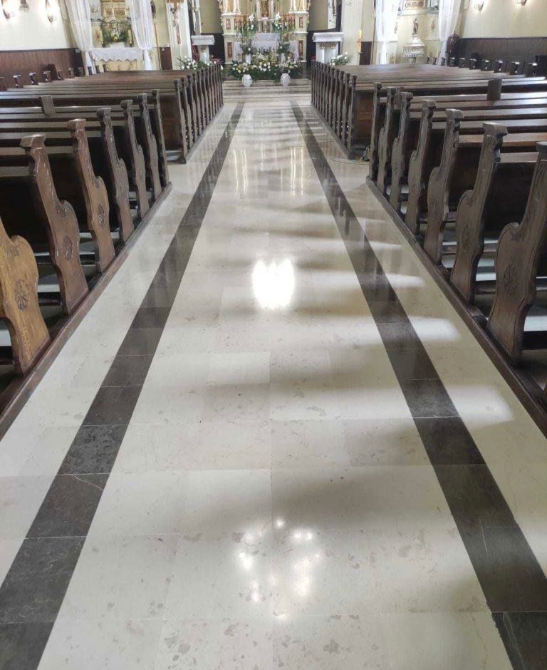 Biało-szara, wypolerowana posadzka z kamiennych płyt w kościele, z rzędami drewnianych ławek po bokach, prowadząca do ołtarza w tle.