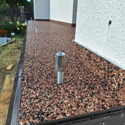 DAMIAN SITEK KAMIENNE DYWANY - Balkon z wykończeniem z drobnych kamyczków w odcieniach brązu i czerni, widoczny śrubunek montażowy, elewacja budynku w tle.