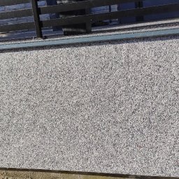 DAMIAN SITEK KAMIENNE DYWANY - Szara elewacja budynku wykończona tynkiem mozaikowym z widocznym oknem i czarną balustradą na dachu.