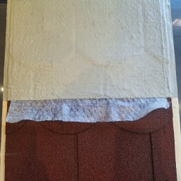 DAMIAN SITEK KAMIENNE DYWANY - Przekrój warstwowy dachu: brązowe gonty bitumiczne, biała membrana hydroizolacyjna i szara warstwa izolacyjna, prezentacja materiałów.