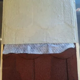 Przekr&oacute;j warstwowy dachu: brązowe gonty bitumiczne, biała membrana hydroizolacyjna i szara warstwa izolacyjna, prezentacja materiał&oacute;w.