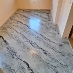 Jasna posadzka w salonie, imitująca marmur, z delikatnym połyskiem. Widoczne ściany w odcieniach beżu i okno z białymi ramami. Nowoczesny design podłogi.