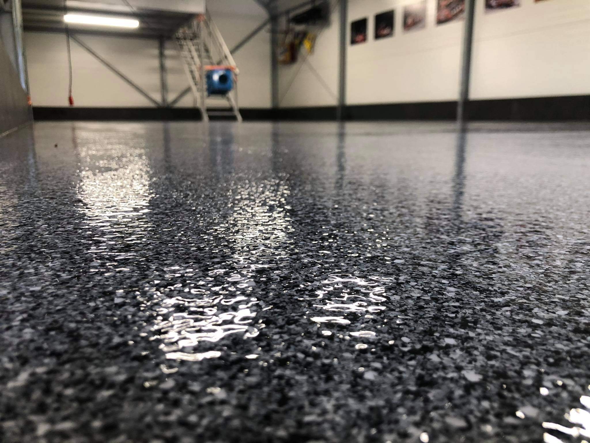 Błyszcząca, ciemna posadzka z drobnymi, jasnymi inkluzjami, odbijająca światło lamp sufitowych w hali przemysłowej. W tle widoczny metalowy stelaż i drabina.