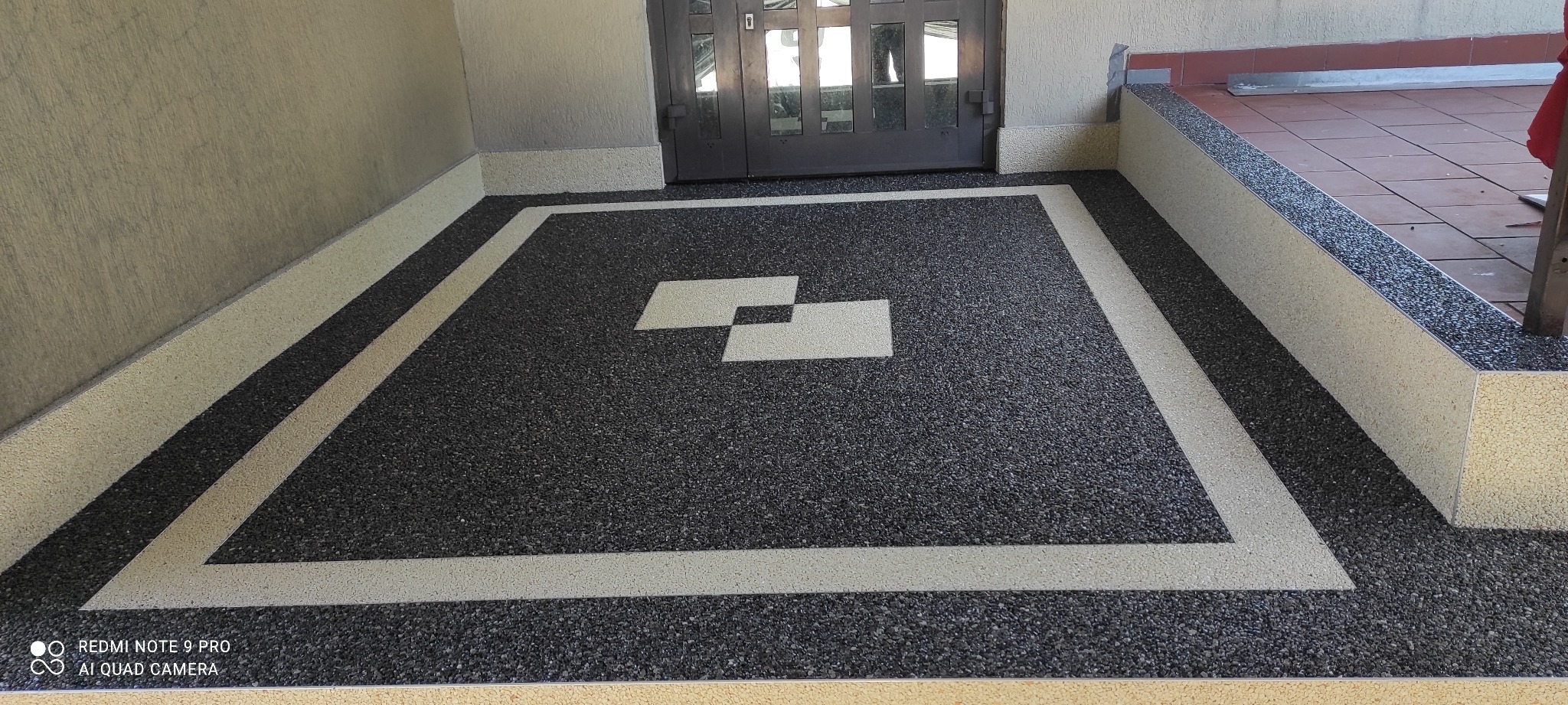 Wykończona posadzka z ciemnego kamyczka z jasnymi obwódkami i geometrycznym wzorem w centrum, zrealizowana w Julianowie, widoczna przed wejściem do budynku.