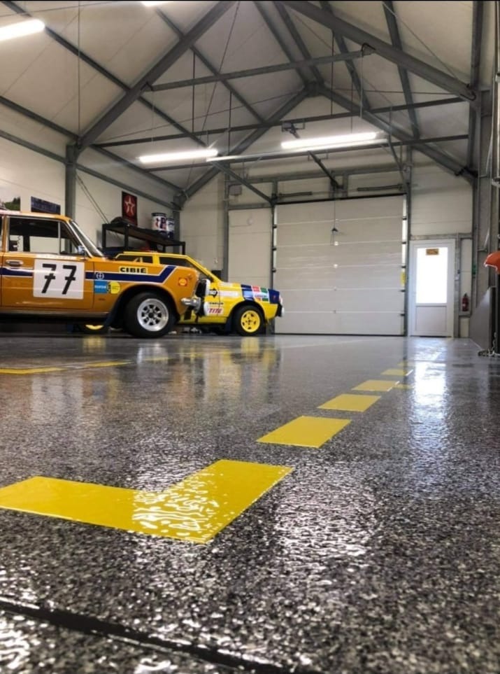 Błyszcząca, szara posadzka w garażu z żółtymi, kwadratowymi oznaczeniami, na której stoi żółty samochód rajdowy z numerem 77.