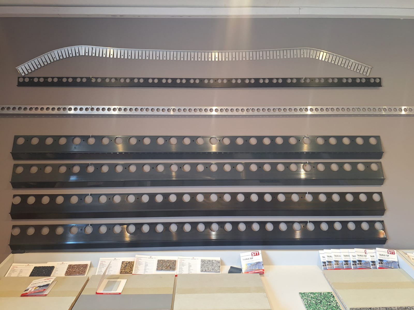 Ekspozycja profili montażowych z otworami i perforacjami, różne modele i kolory na szarym tle, próbki materiałów budowlanych na białym blacie poniżej.