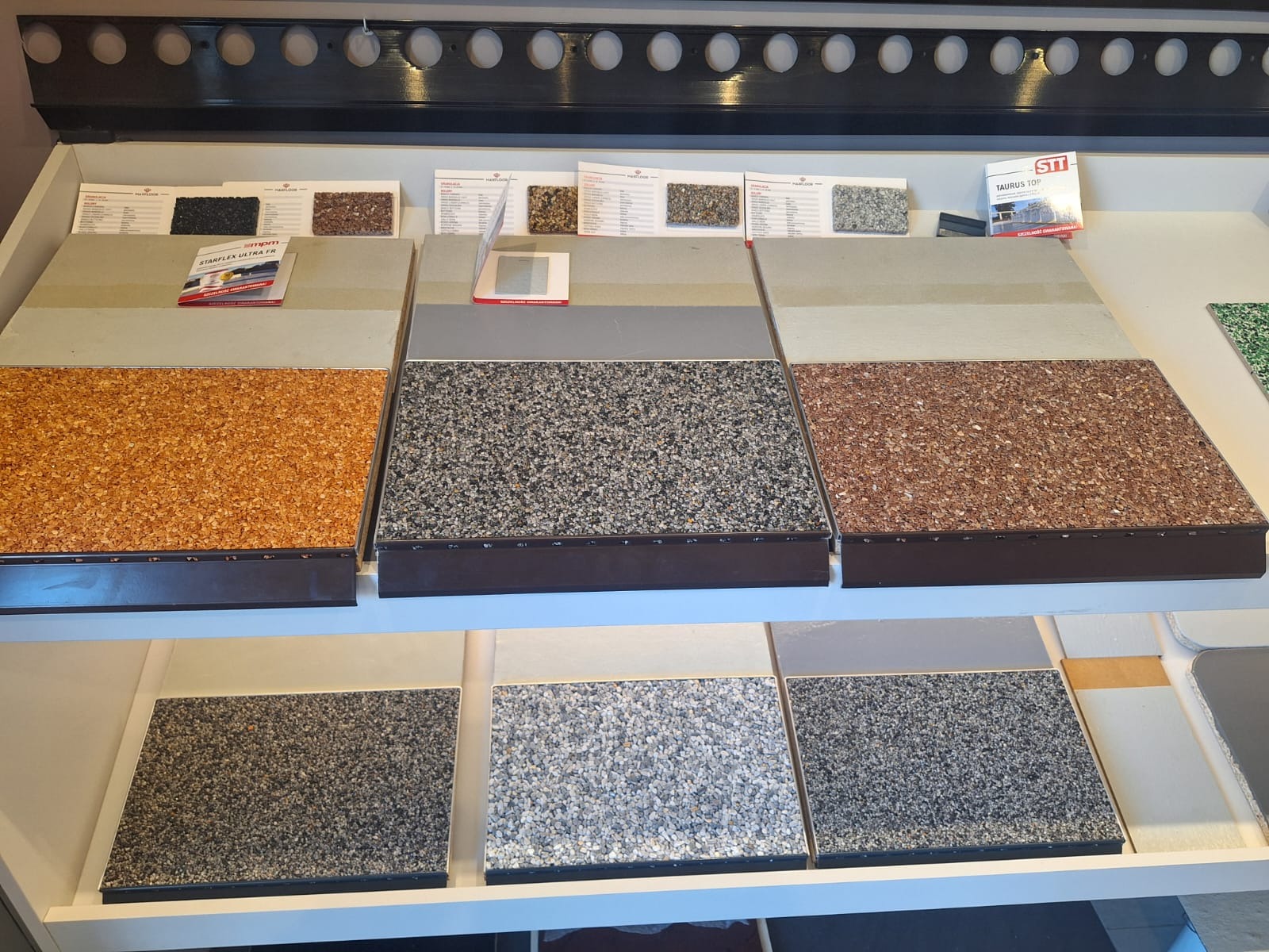 Ekspozycja próbek wielowarstwowych posadzek z różnymi kolorami i fakturami kruszywa, prezentowane na półkach w salonie z katalogami producentów.