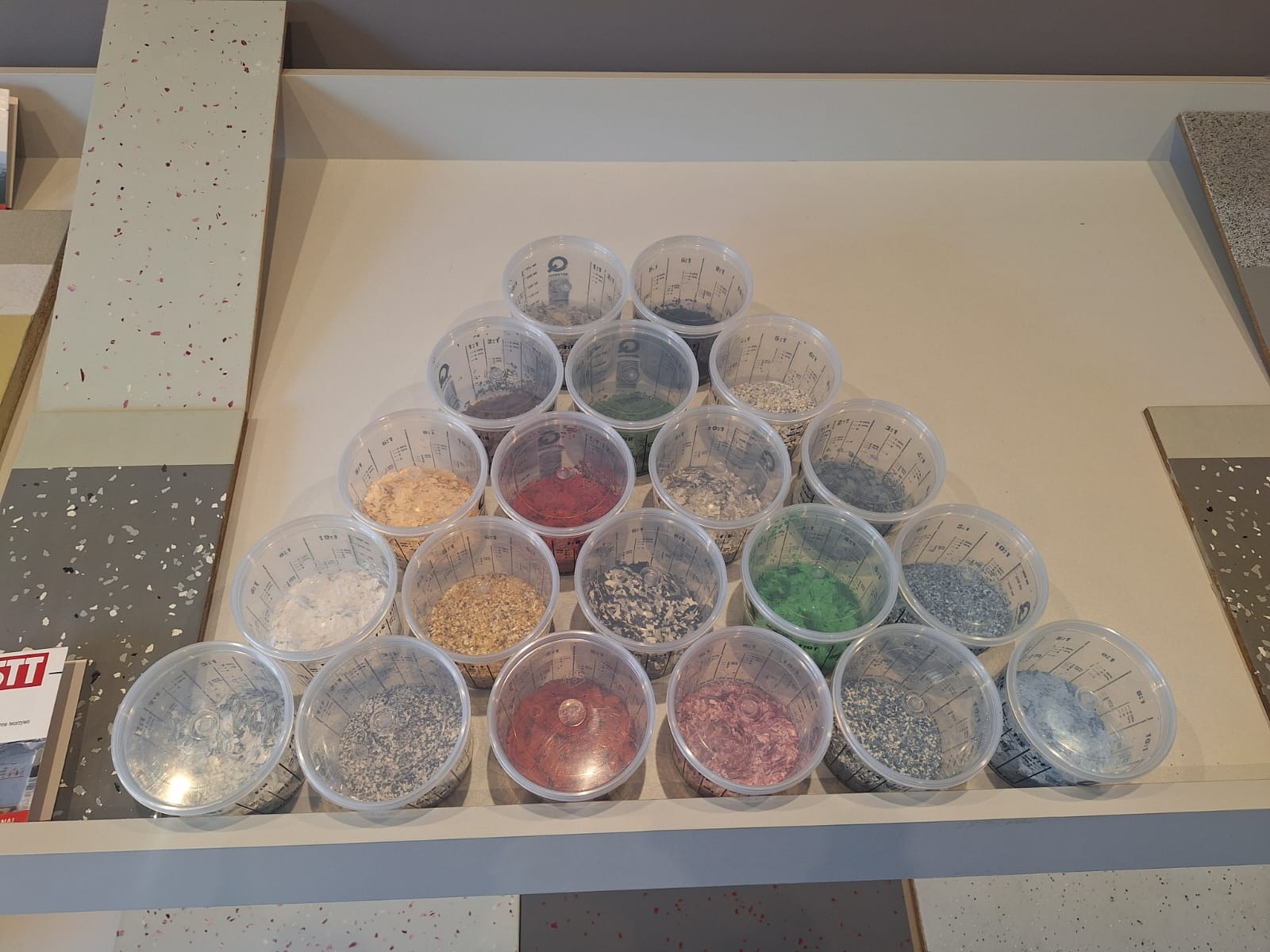 Prezentacja próbek żywicy epoksydowej w przezroczystych pojemnikach, ułożonych w piramidę na blacie w Julianowie, obok paneli z gotowymi posadzkami żywicznymi w różnych kolorach i wzorach.