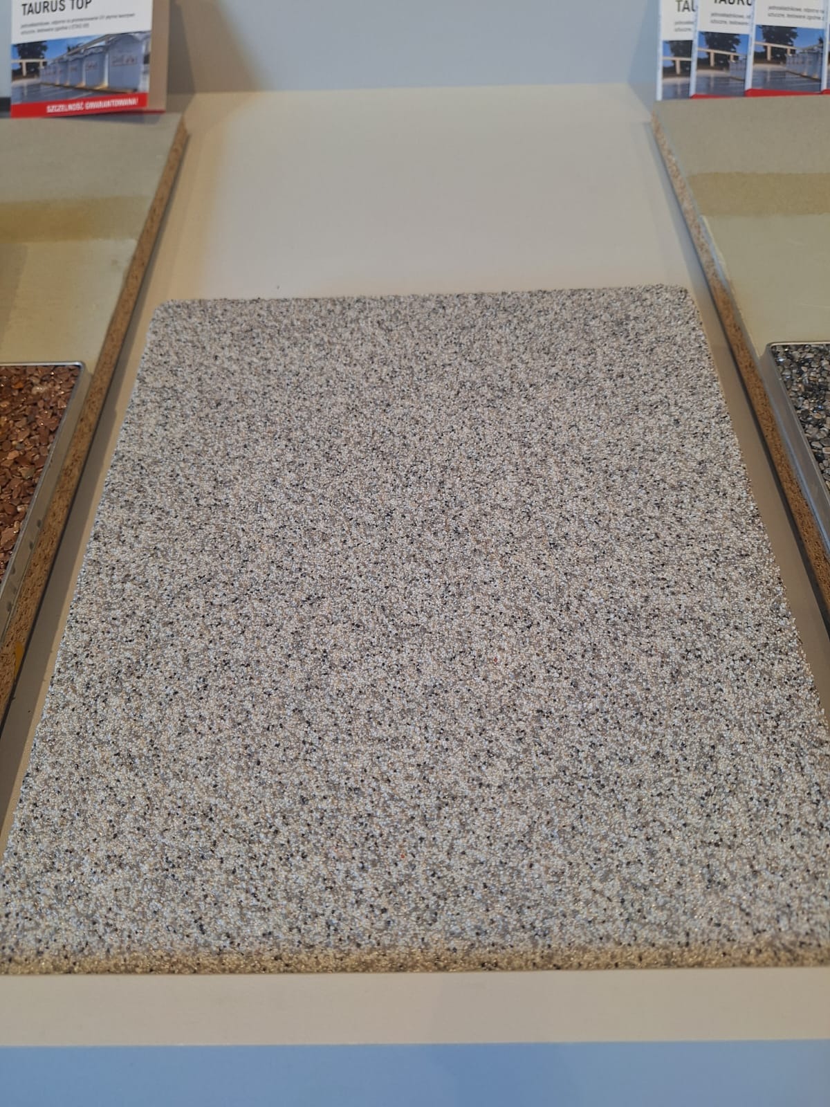 Próbka szarej posadzki z drobnym kruszywem, prezentowana obok innych próbek na ekspozycji.