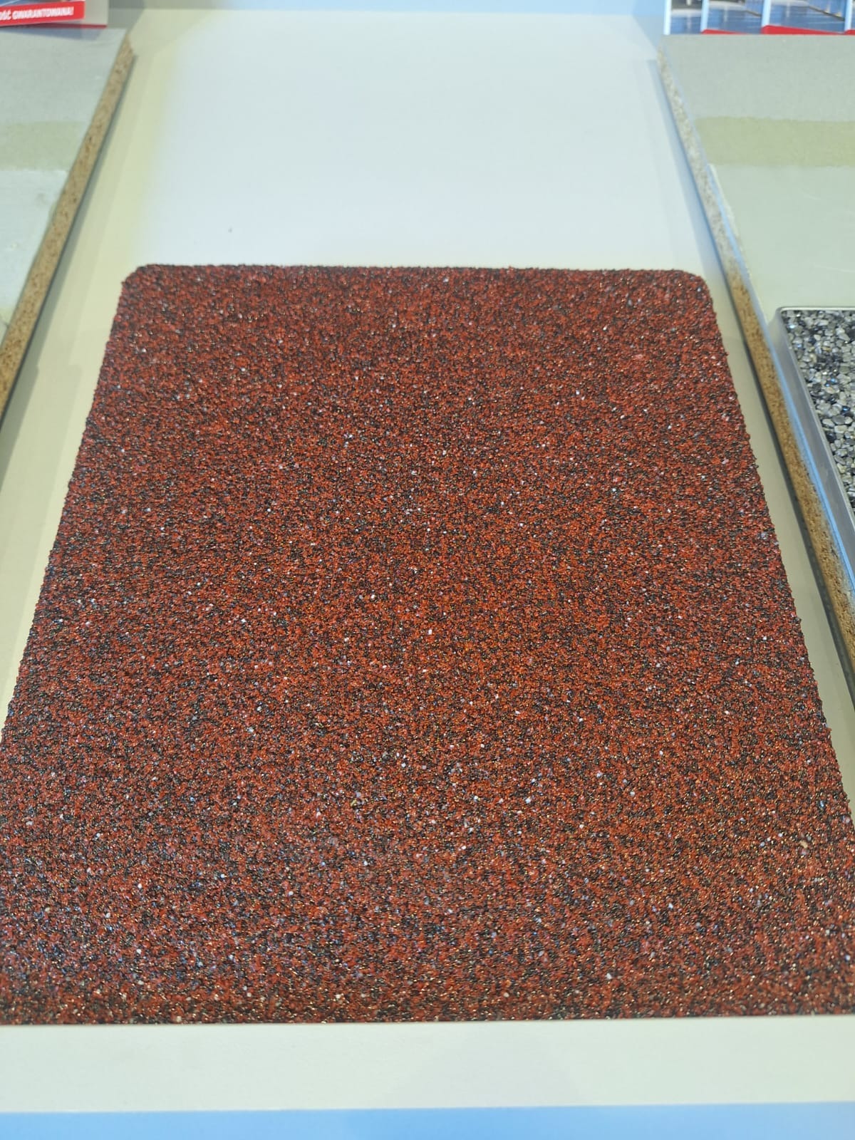 Próbka posadzki o czerwono-brązowej, ziarnistej fakturze, otoczona ramkami z innych materiałów wykończeniowych.