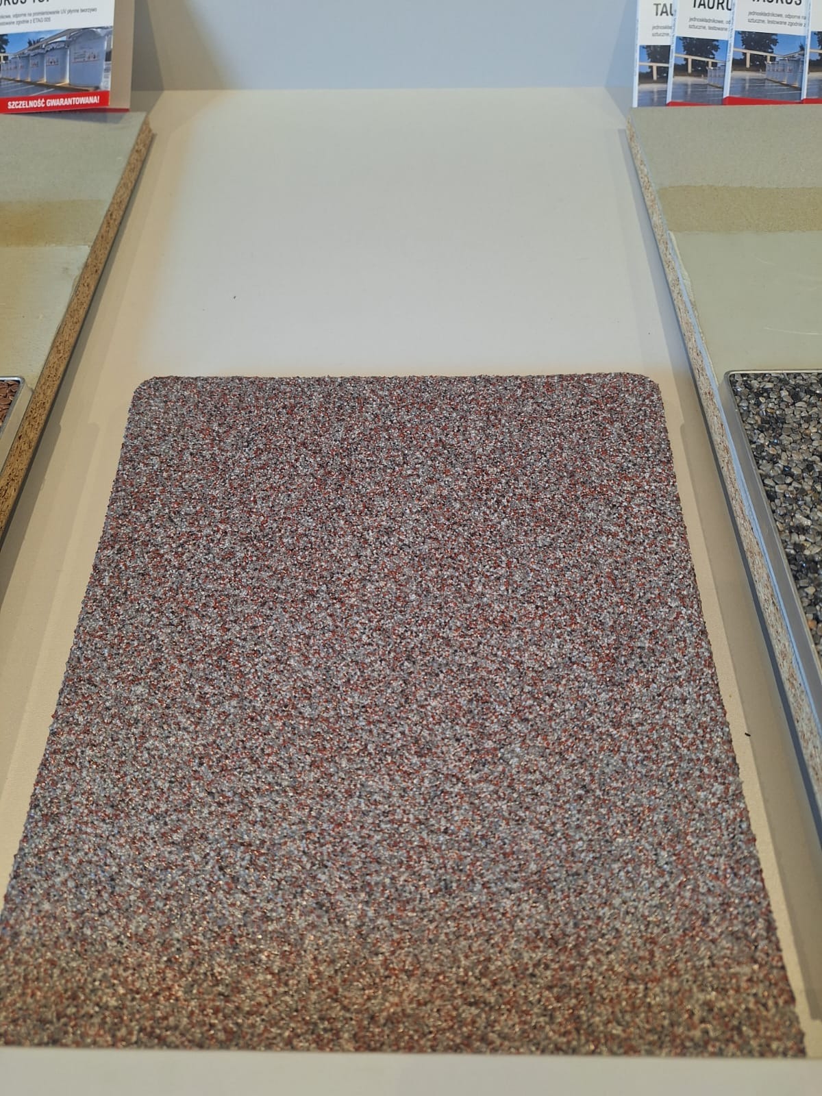 Próbka posadzki żywicznej w kolorze czerwono-szarym, drobnoziarnista tekstura, prezentowana na ekspozycji w salonie, widoczne inne próbki podłóg w tle.
