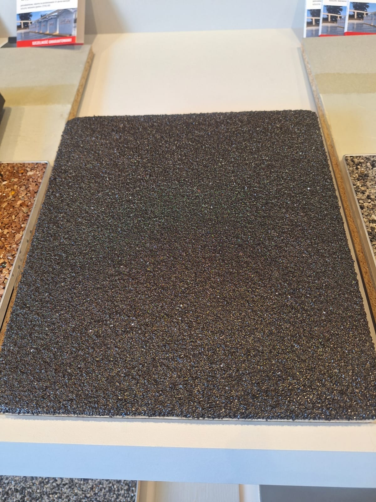 Próbka ciemnoszarej posadzki o drobnej ziarnistej strukturze, otoczona innymi materiałami wykończeniowymi na jasnym blacie.