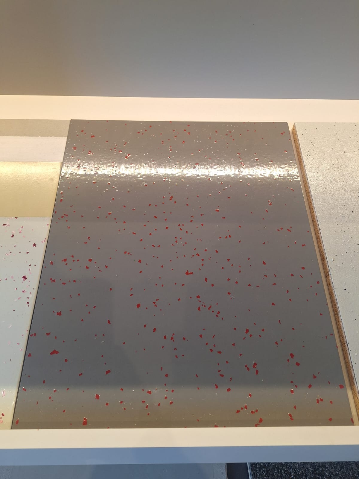 Próbka szarej, błyszczącej posadzki z czerwonymi, nieregularnymi drobinkami na ekspozytorze z innymi wariantami kolorystycznymi.