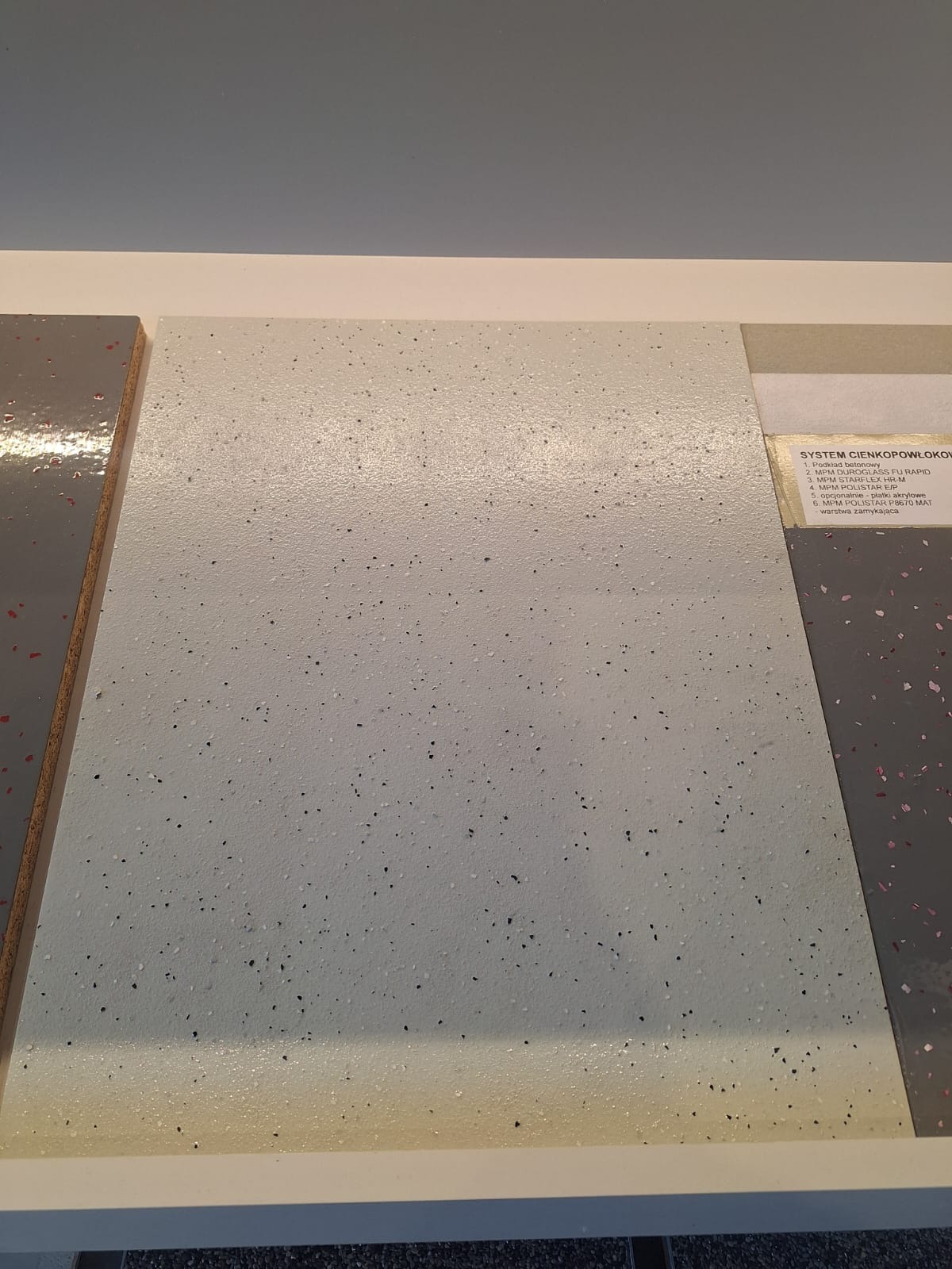 Próbki różnych wykończeń podłogowych, w tym jasnoszara powierzchnia z drobnymi, czarnymi i szarymi plamkami, oraz etykieta opisująca system cienkopowłokowy z wykazem składników i warstw.