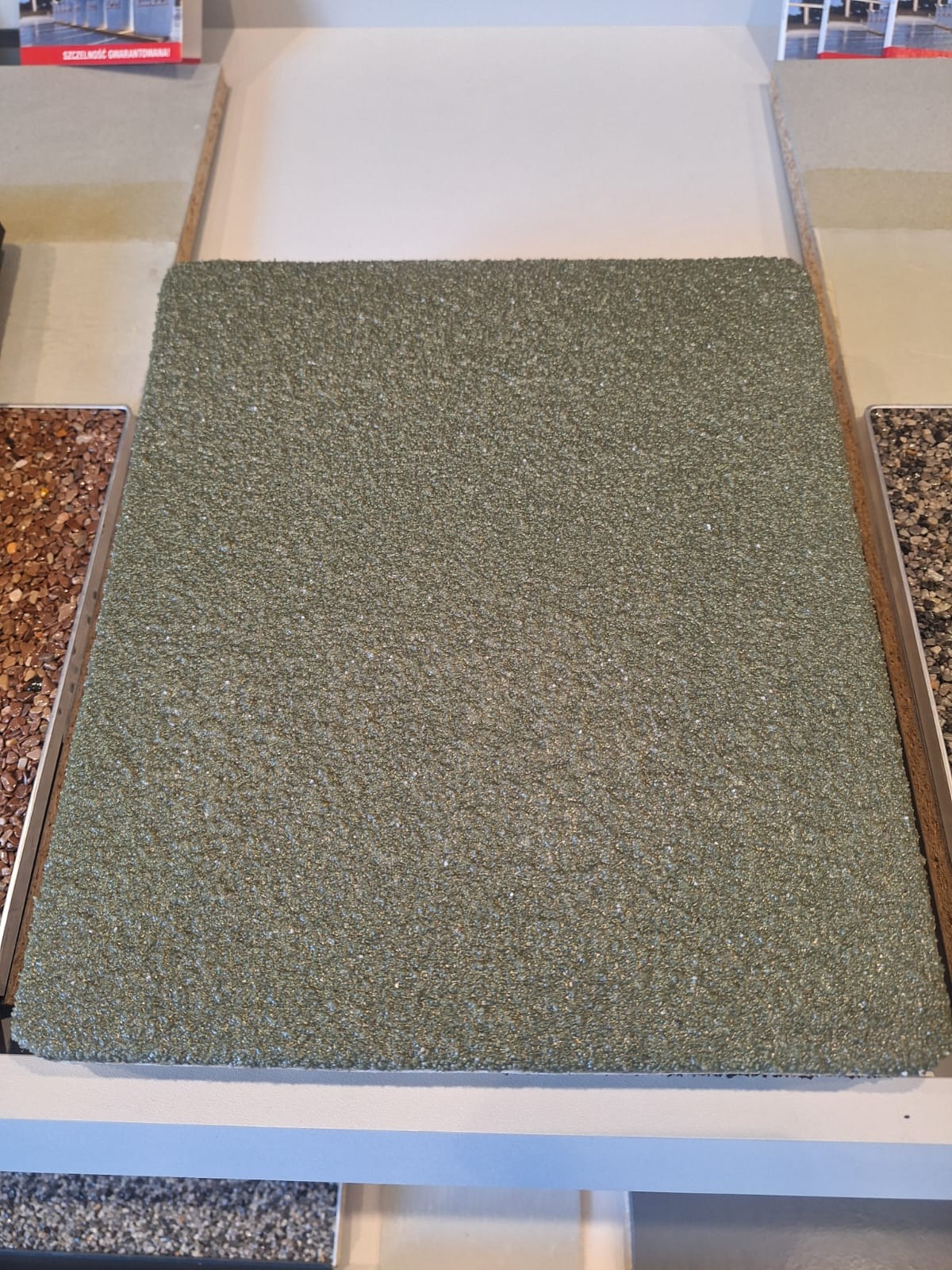 Próbka zielonej posadzki o drobnej, chropowatej fakturze, otoczona innymi materiałami wykończeniowymi na ekspozytorze.