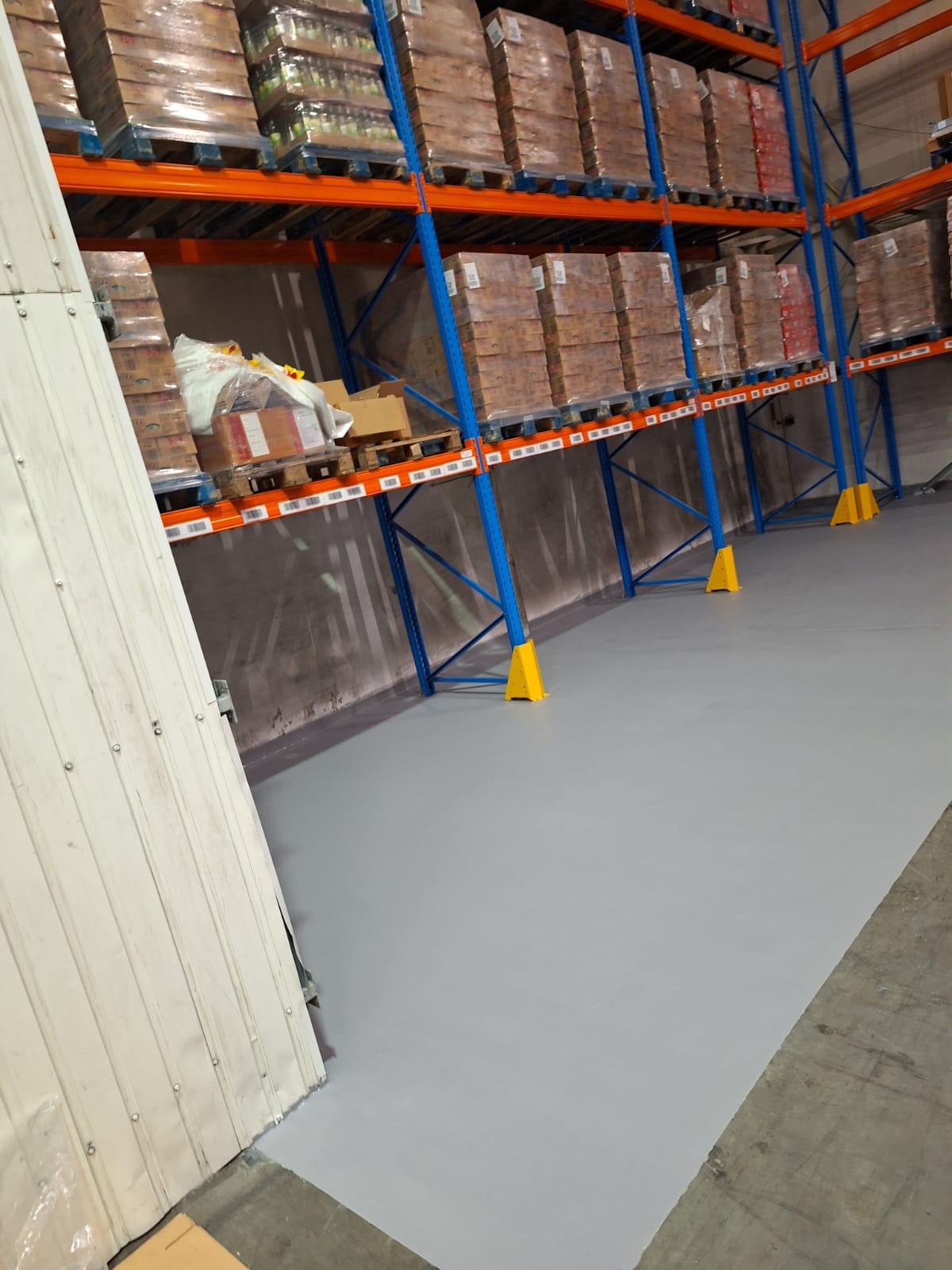 Magazyn z regałami i szarą, gładką posadzką żywiczną. Widoczne żółte zabezpieczenia regałów. Ściana w tle. Ujęcie z dołu, podkreślające powierzchnię podłogi.