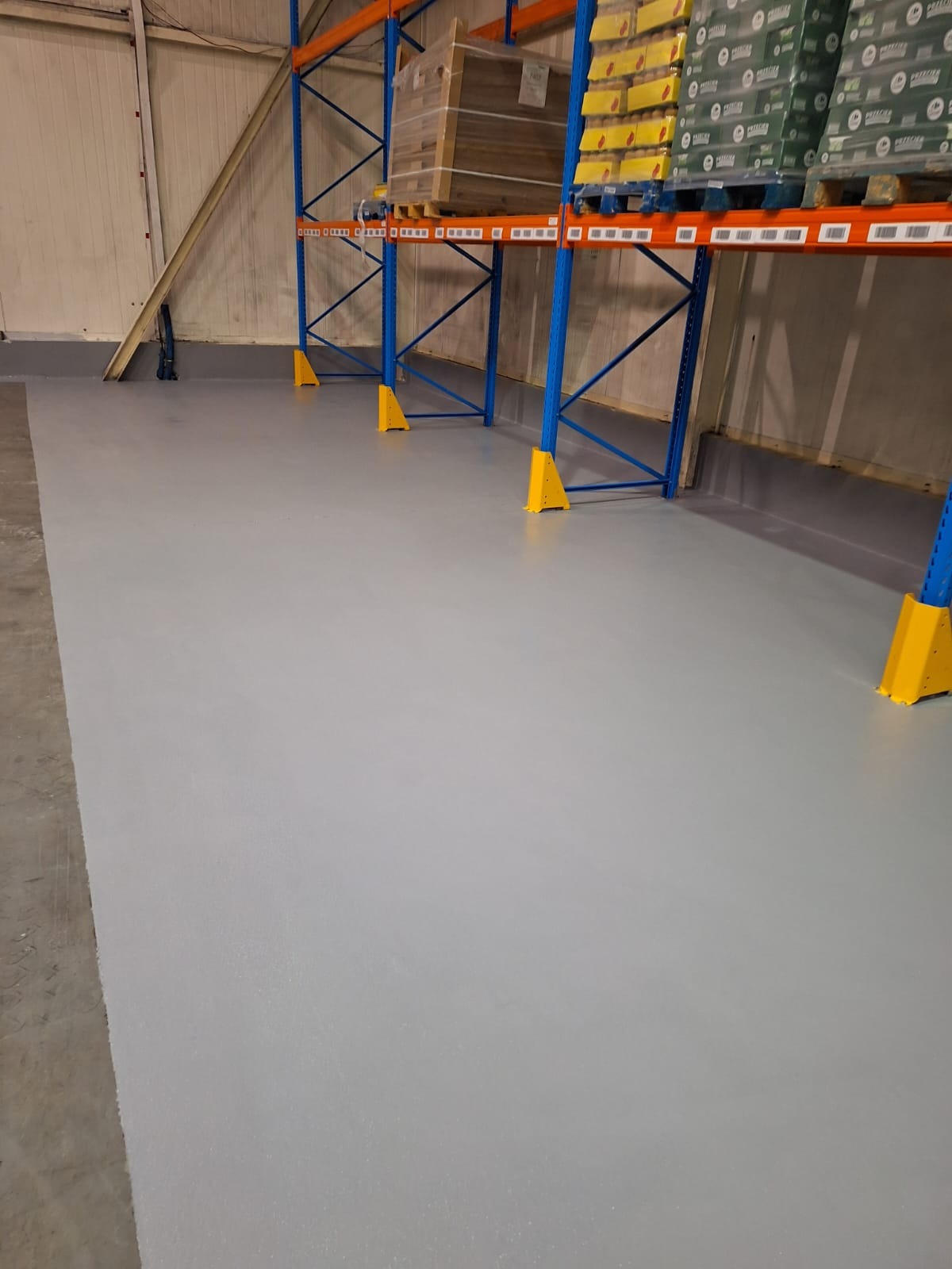 Szara, gładka posadzka w hali magazynowej, z fragmentem regałów w tle. Widoczne żółte osłony nóg regałów. Nowa, równa powierzchnia.
