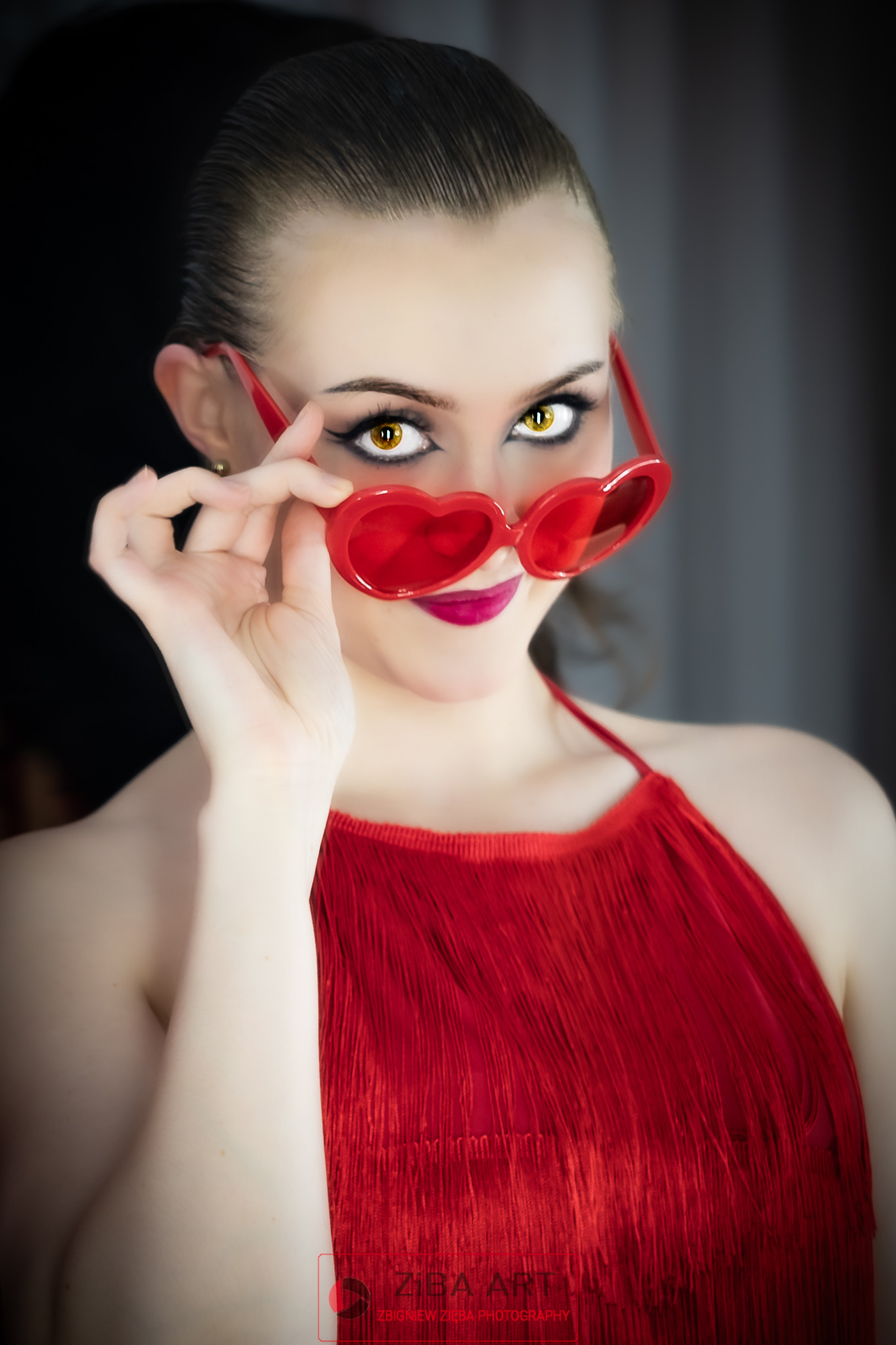Portret kobiety z gładko zaczesanymi włosami, w czerwonej sukience z frędzlami i okularach w kształcie serc, z wyrazistym makijażem oczu i ust.