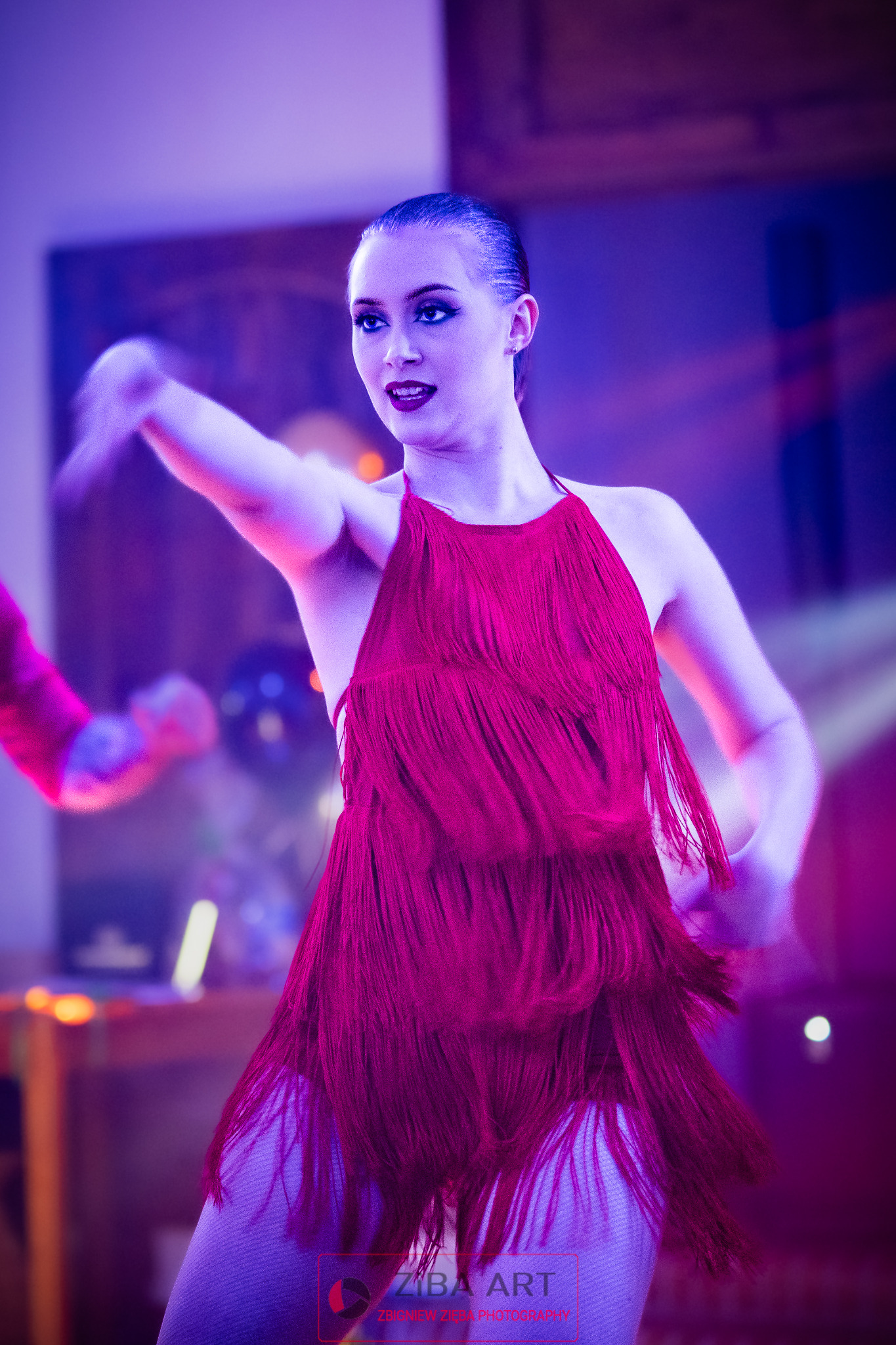 Kobieta w czerwonej sukience z frędzlami tańczy na scenie oświetlonej fioletowym światłem.