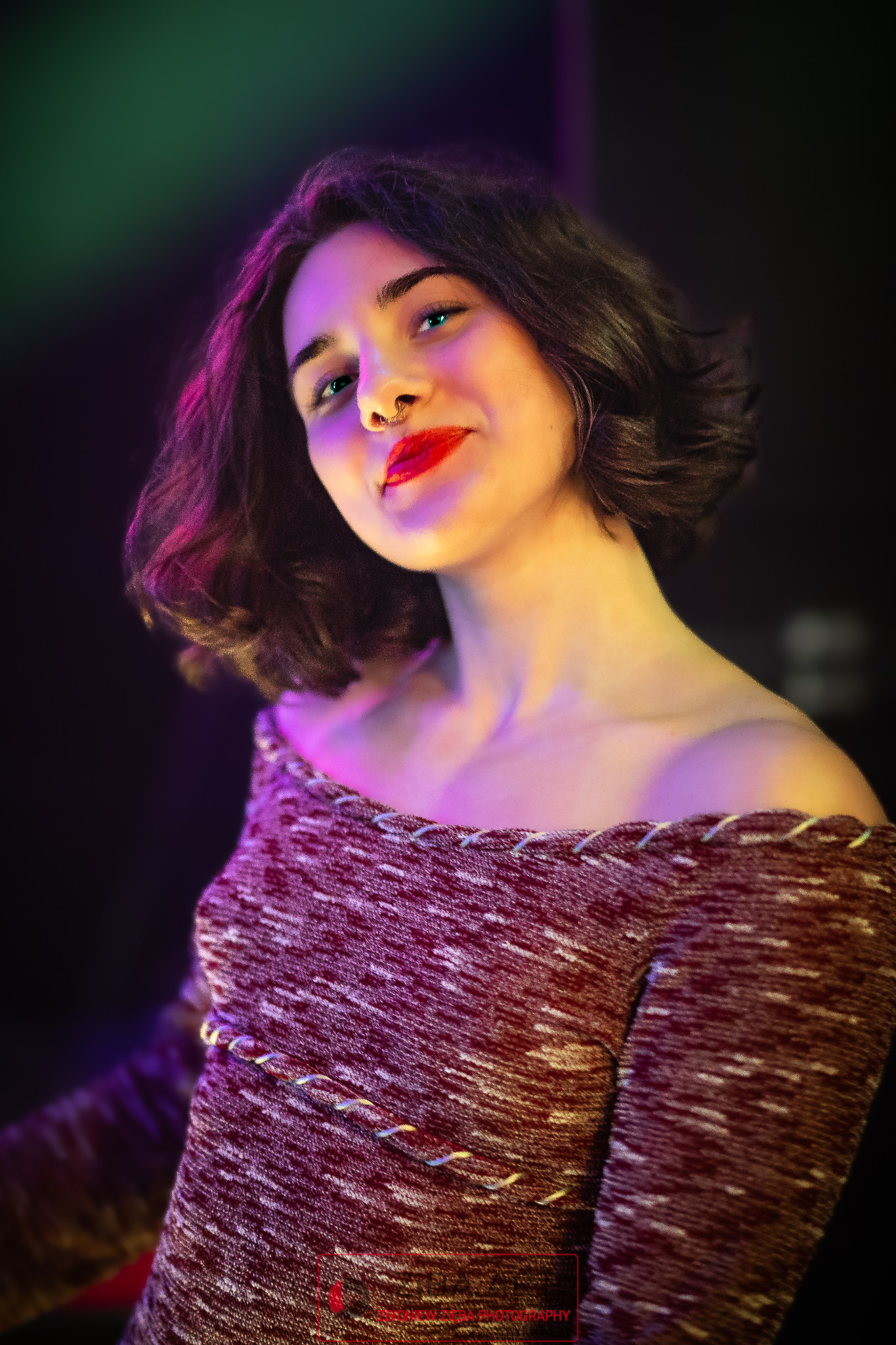 Portret młodej kobiety z piercingiem w nosie, uśmiechniętej, w bluzce z odkrytymi ramionami, oświetlonej kolorowymi światłami (różowe, zielone, fioletowe).